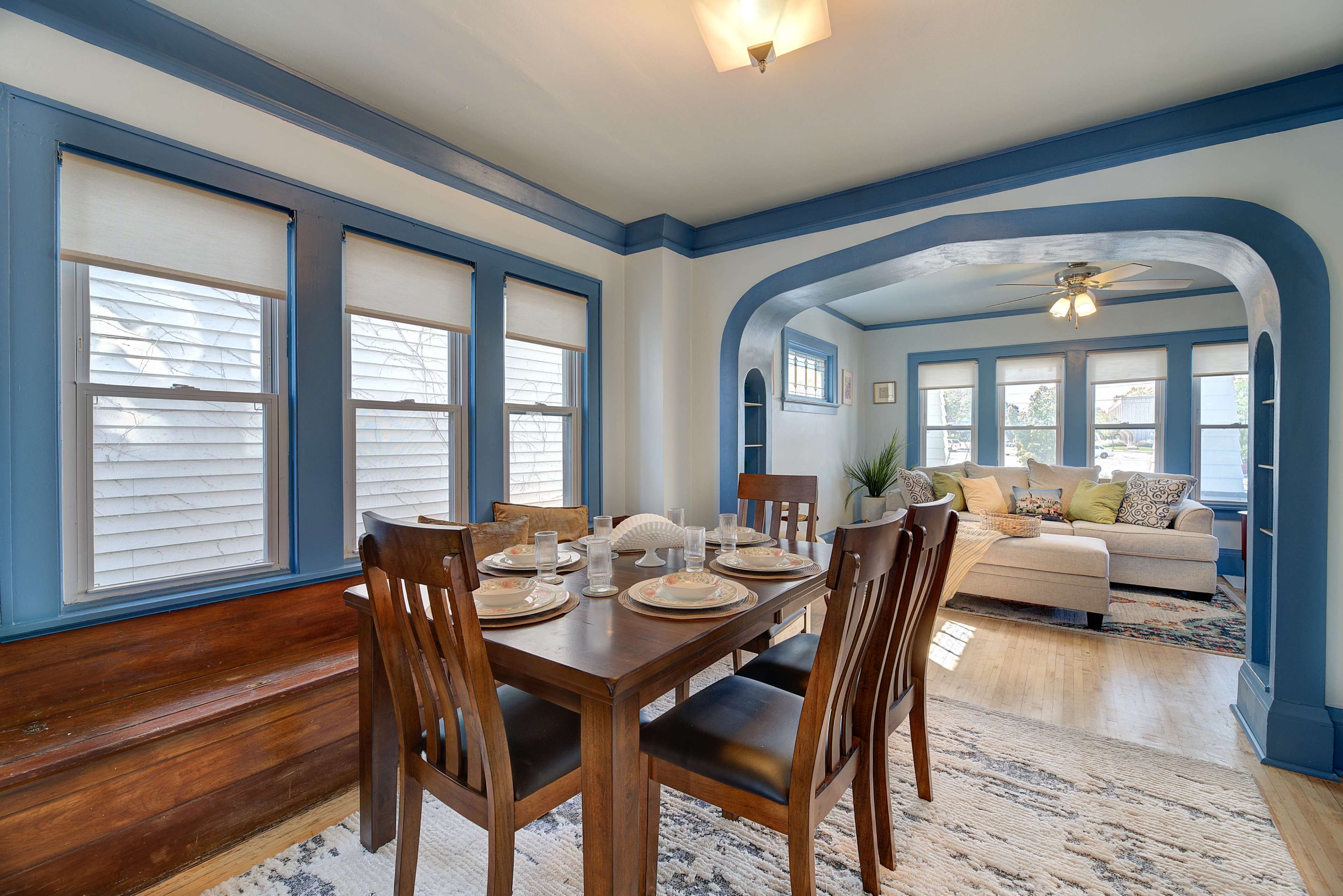 A dining area with a wooden table set for four, adjacent to a living room featuring a light-colored sofa and several windows.