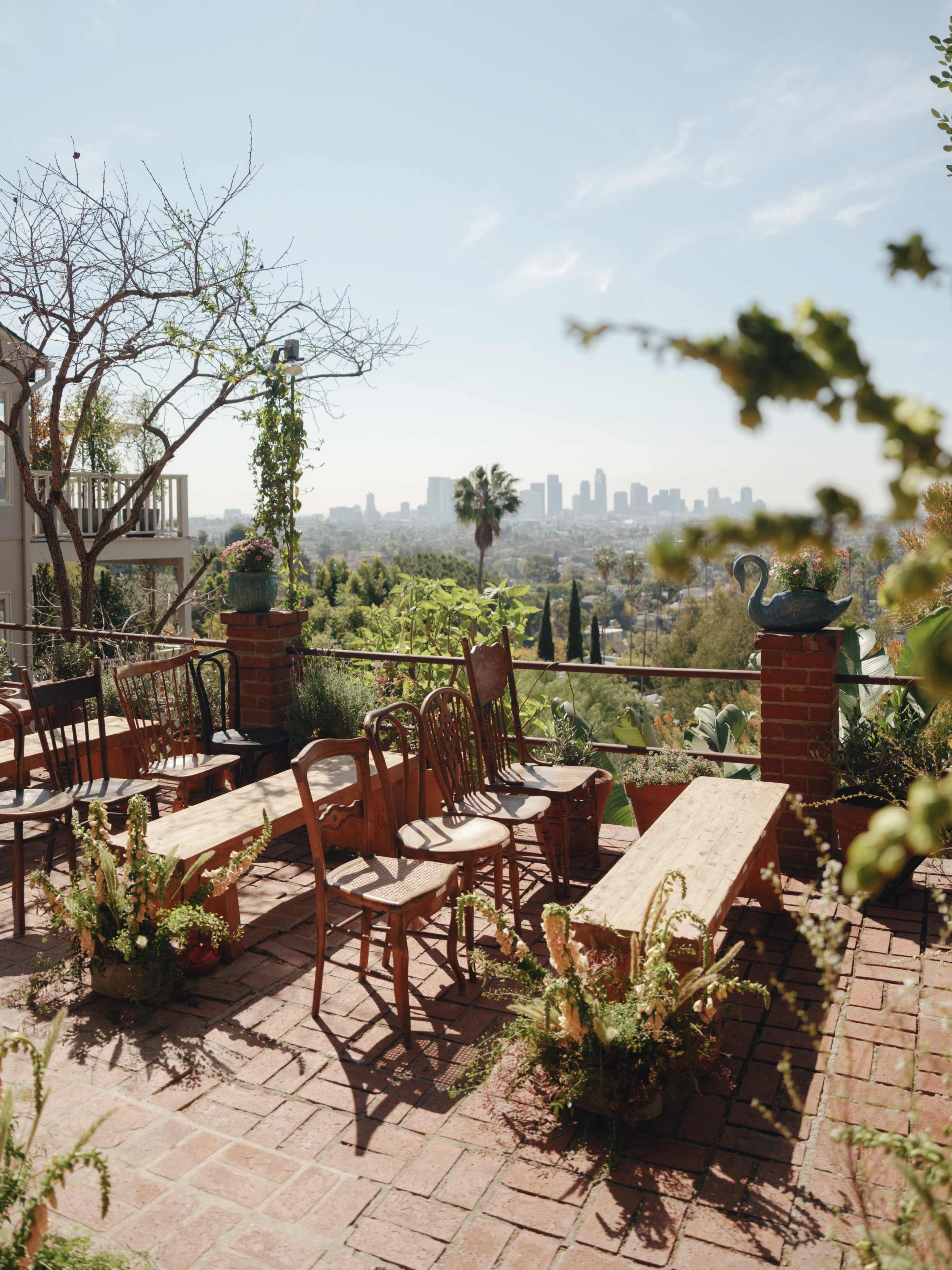 The image shows a terrace with wooden benches and chairs, surrounded by greenery and potted plants, overlooking a city skyline.