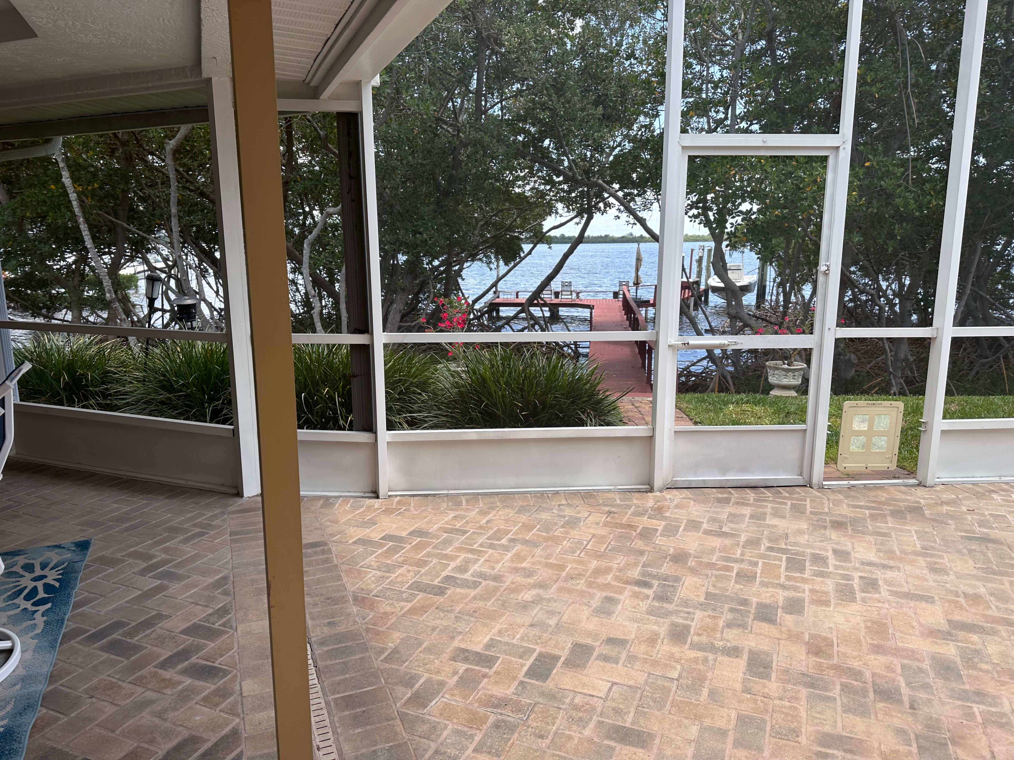 A screened patio overlooks a waterfront with a wooden dock and greenery in the background.