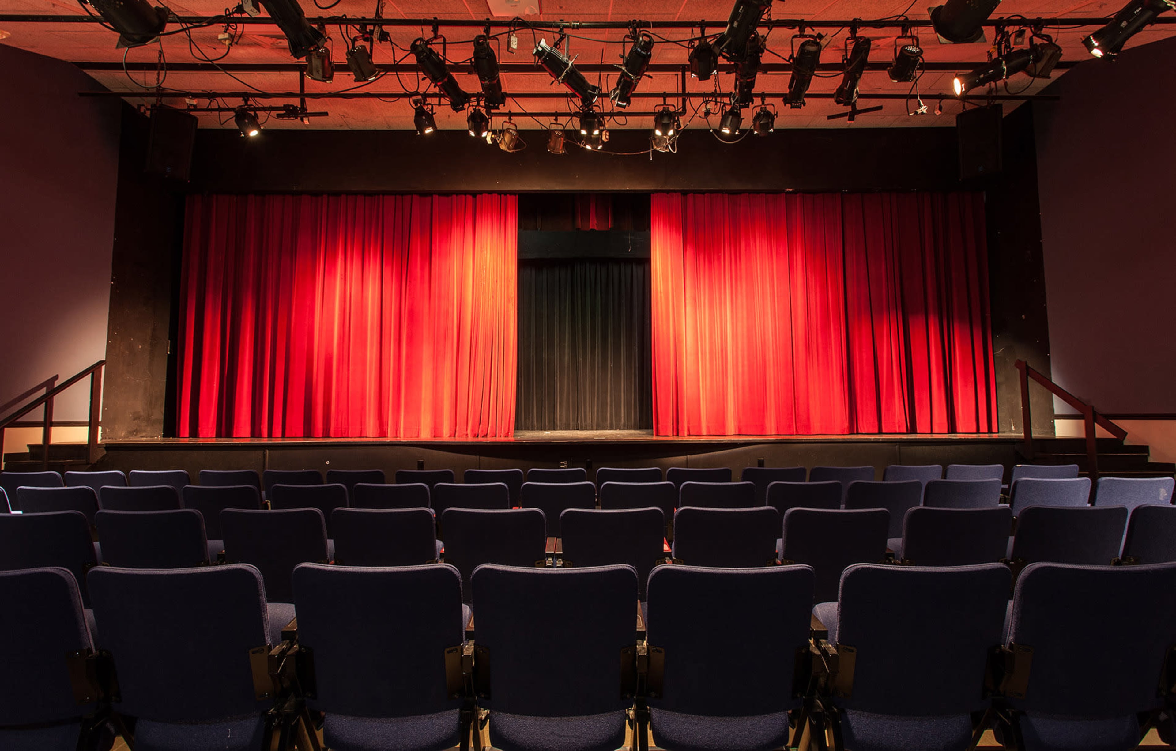 A theater stage with red curtains and a row of empty blue seats facing it.