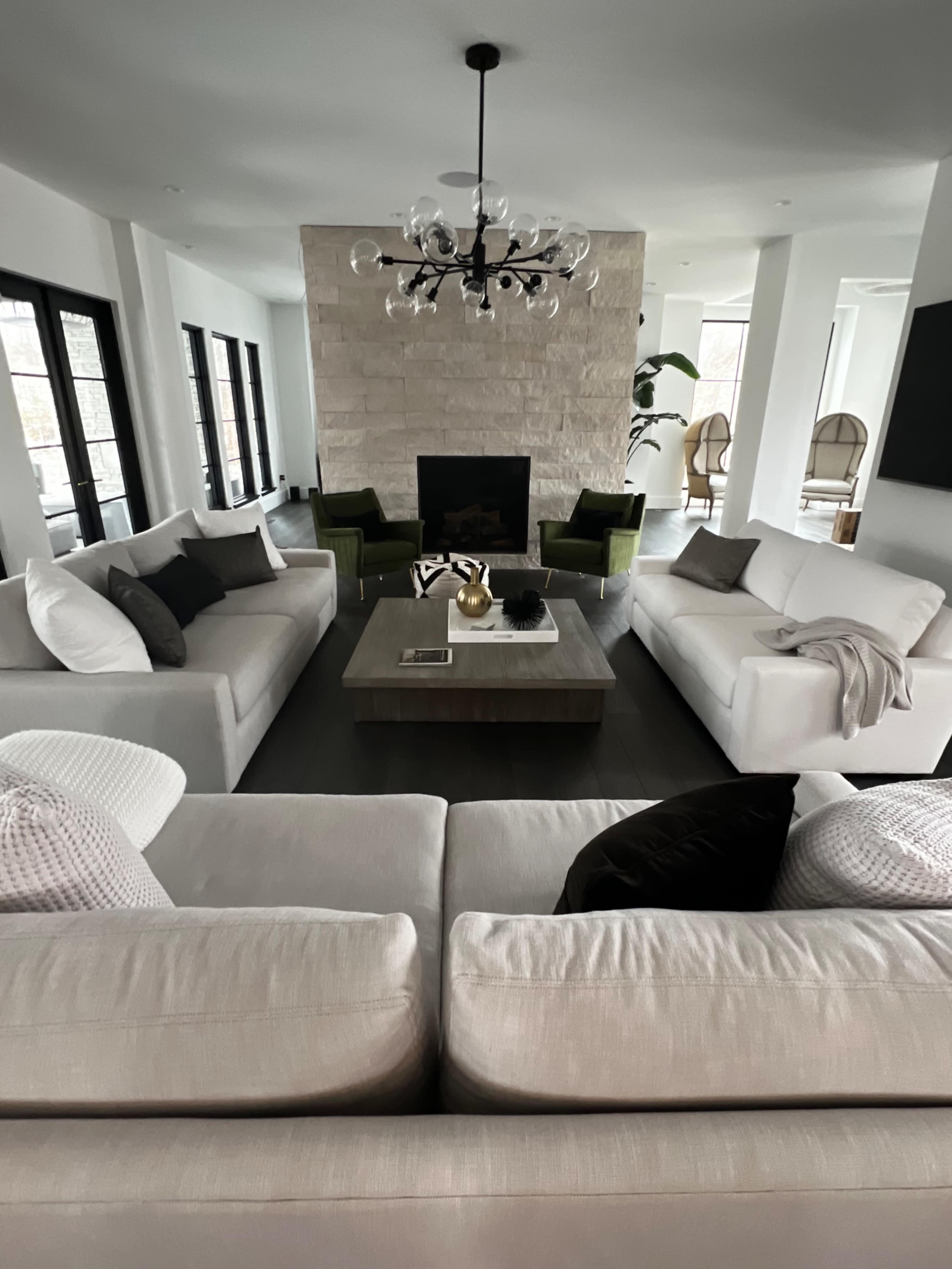 A modern living room with two white sofas facing a wooden coffee table, surrounded by green accent chairs and a stone fireplace.