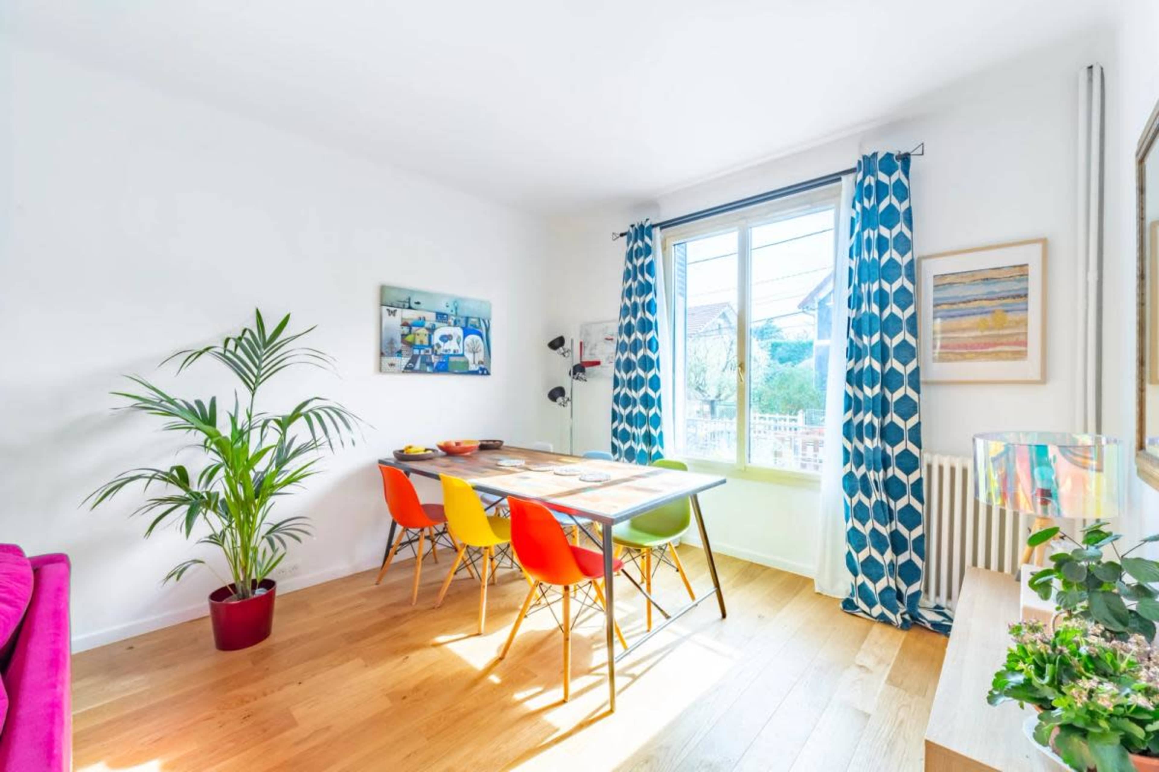 A dining area features a table surrounded by colorful chairs, with a large window letting in natural light and a potted plant in the corner.