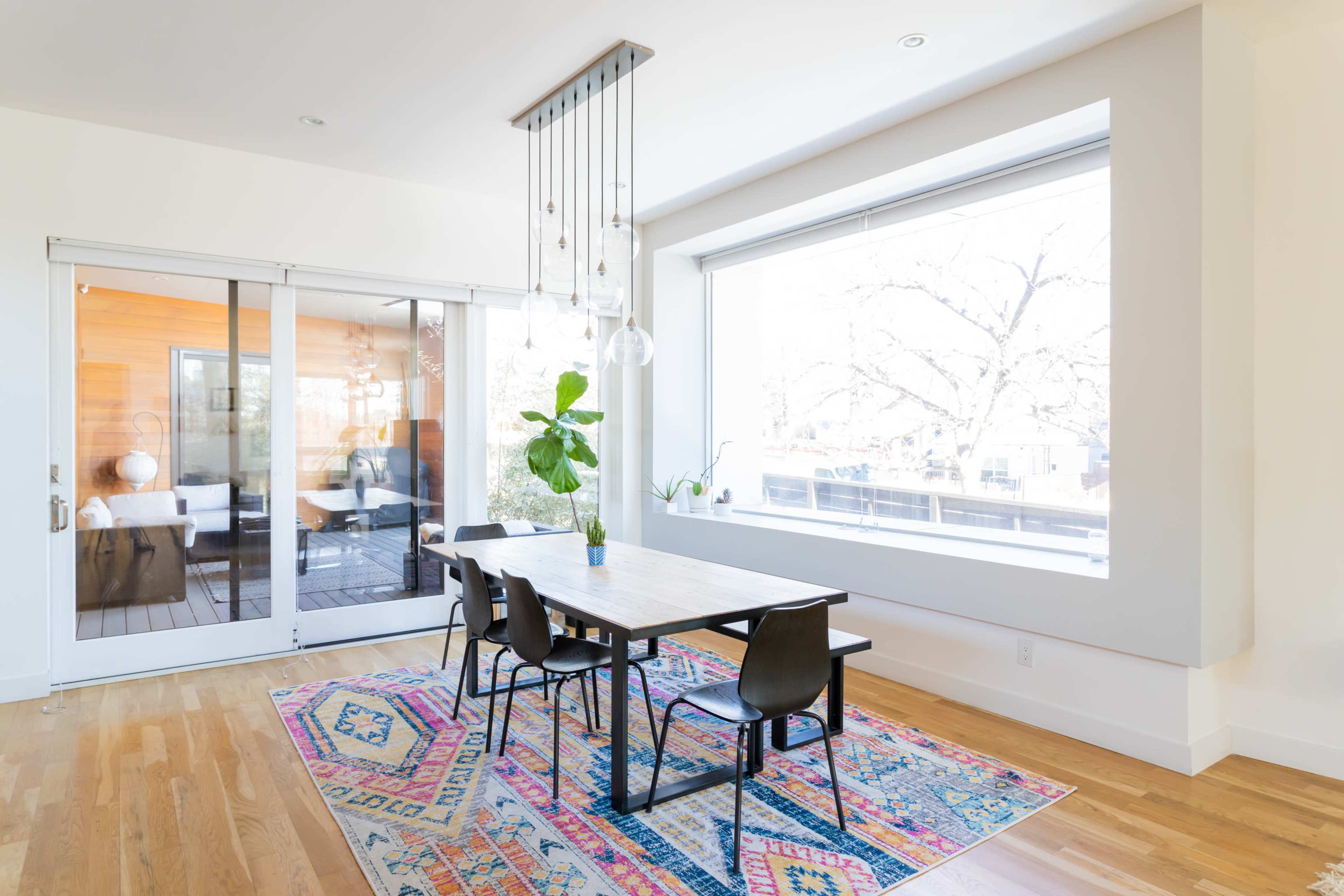 A modern dining area features a wooden table surrounded by black chairs, illuminated by pendant lights, with large windows providing natural light and a view of outdoor trees.