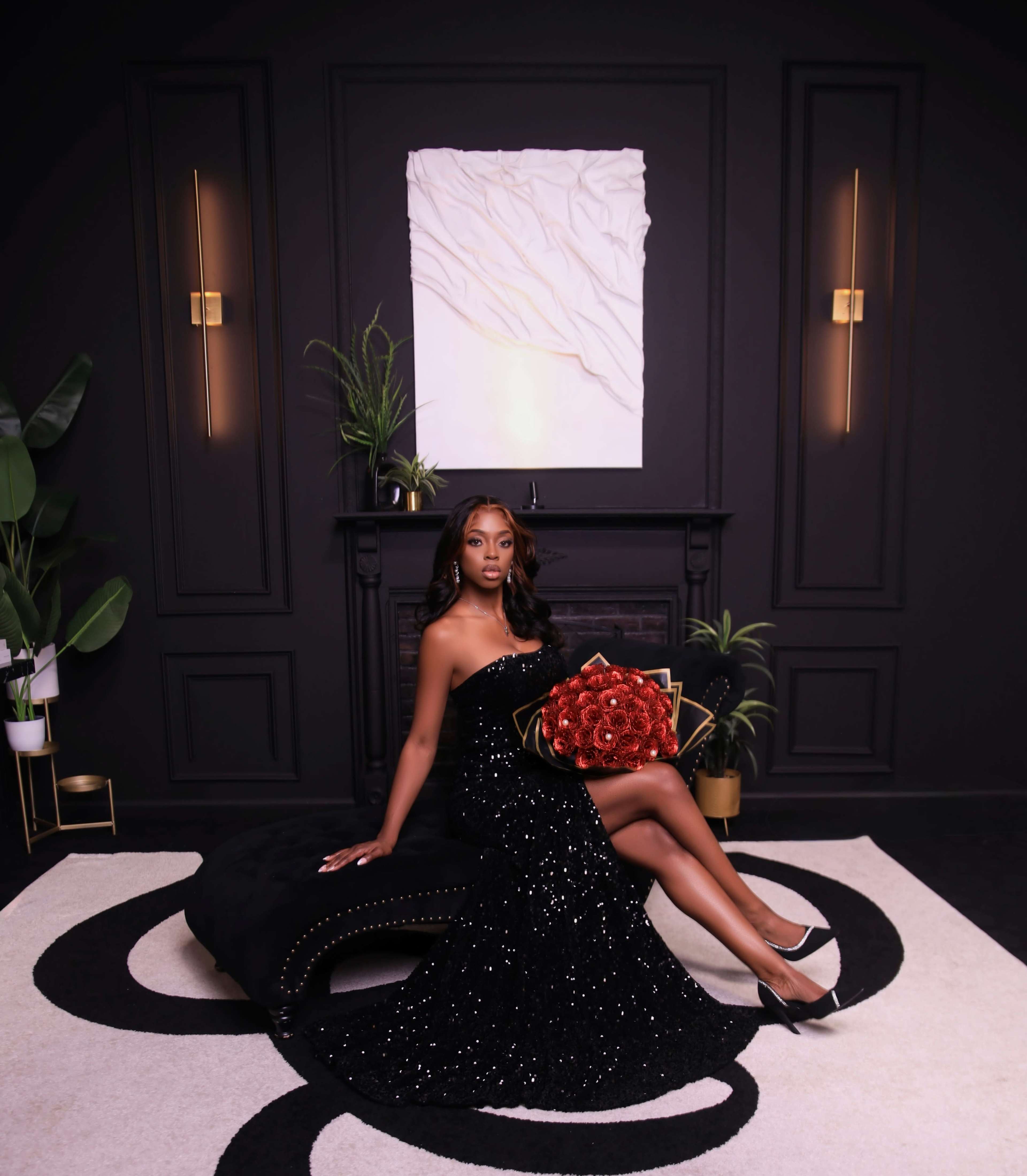A woman in a black sequined gown sits on a plush black chaise, holding a bouquet of red roses in a stylish, dark room with modern decor.
