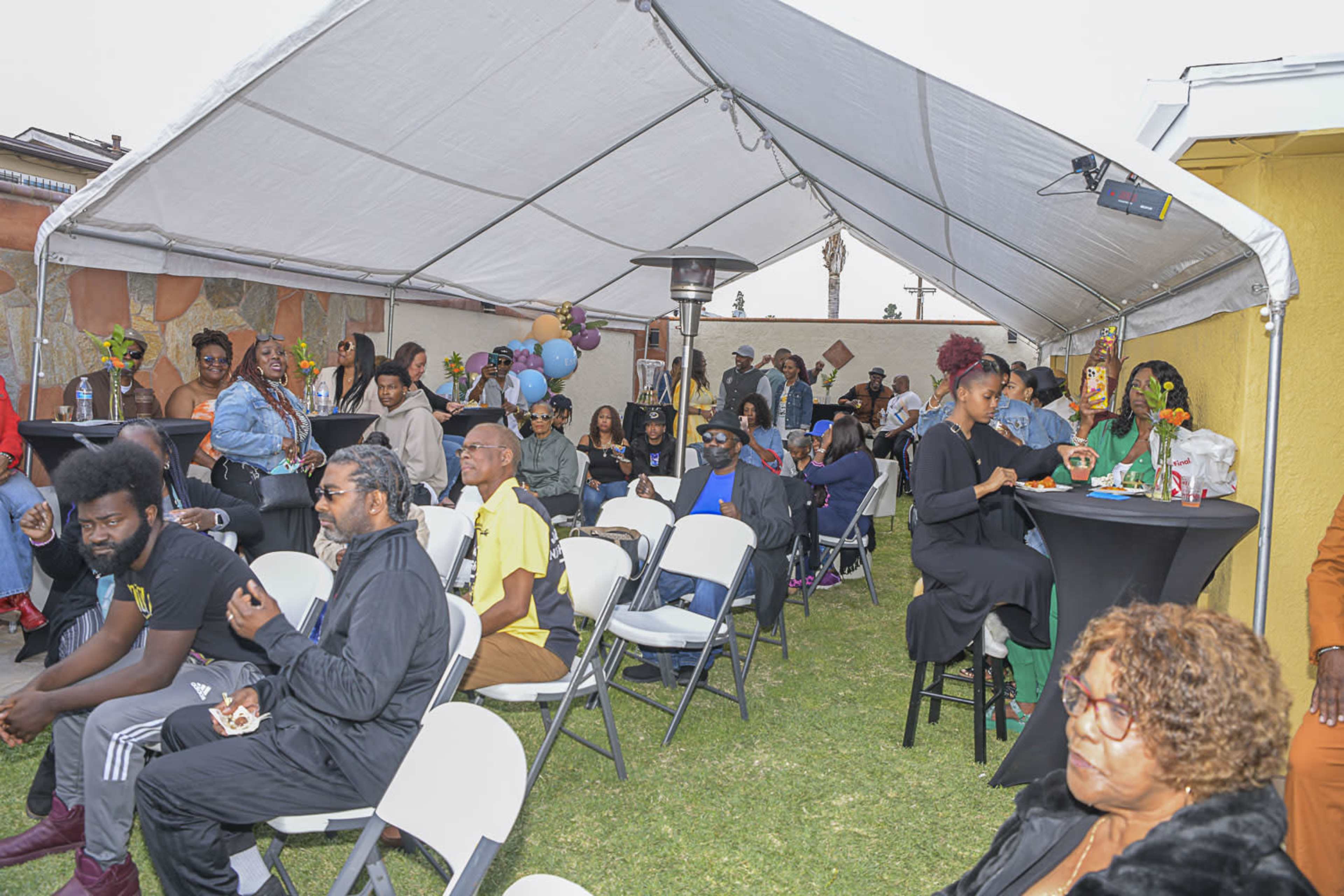A large group of people gathers under a tent at an outdoor event, seated on white chairs and engaging in conversation.