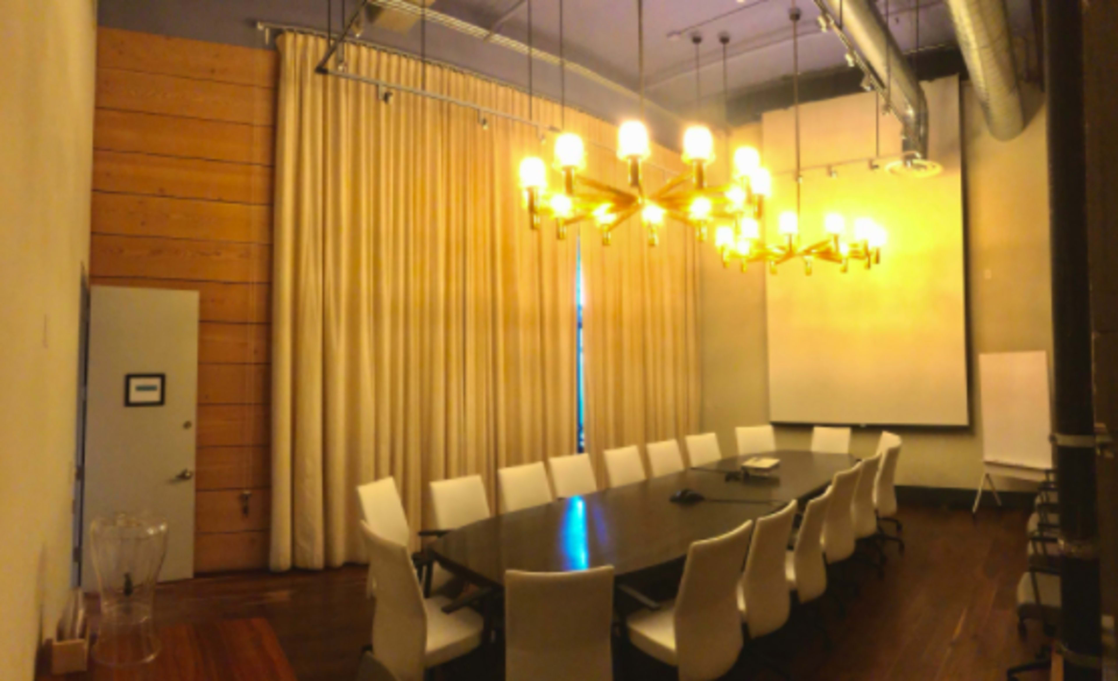A conference room with a long wooden table and white chairs, illuminated by a chandelier, featuring large curtains and a projection screen.