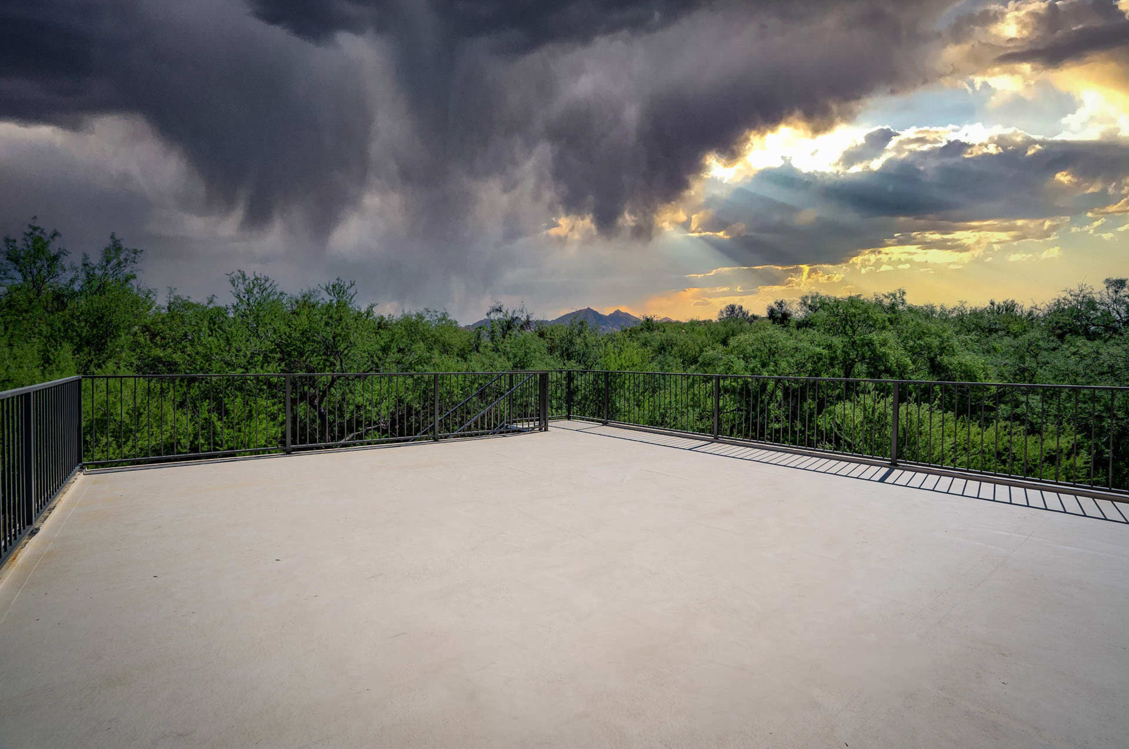 A wide, empty concrete terrace overlooks a dense green landscape under a dramatic sky filled with dark clouds and rays of sunlight breaking through.