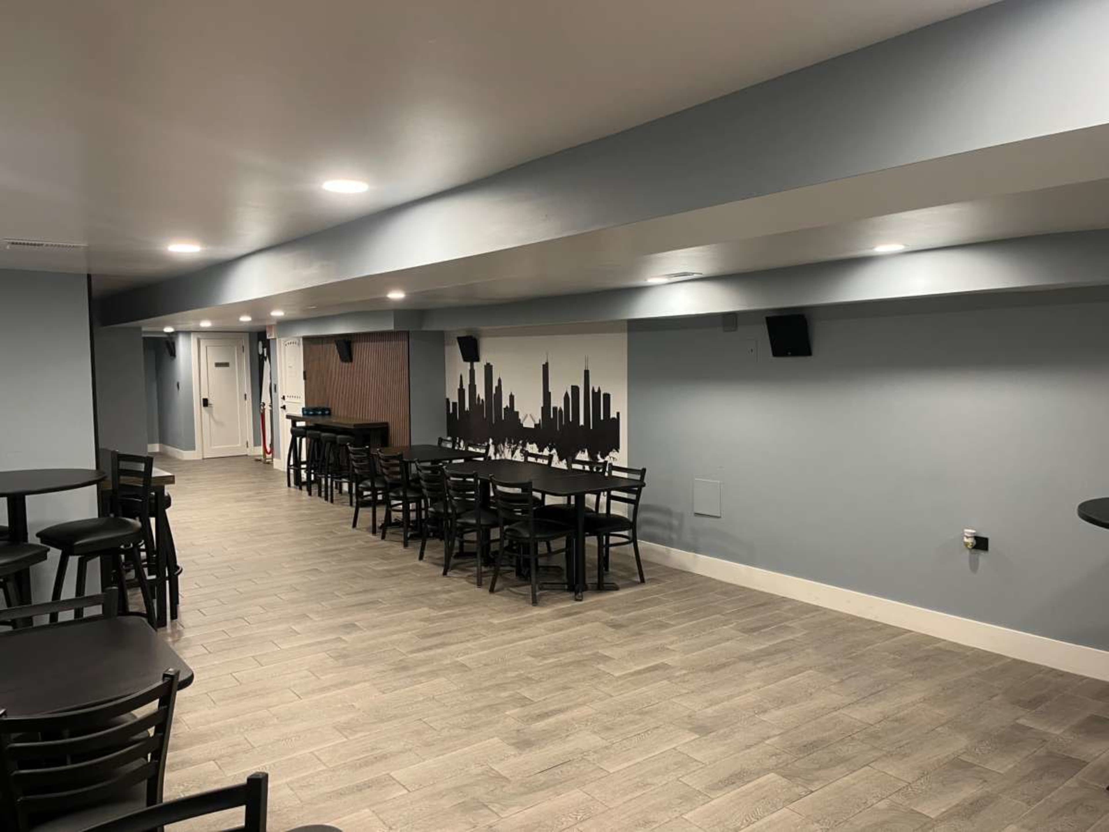 The image shows a dimly lit lounge area featuring several black tables and chairs, with a mural of a city skyline on the wall.