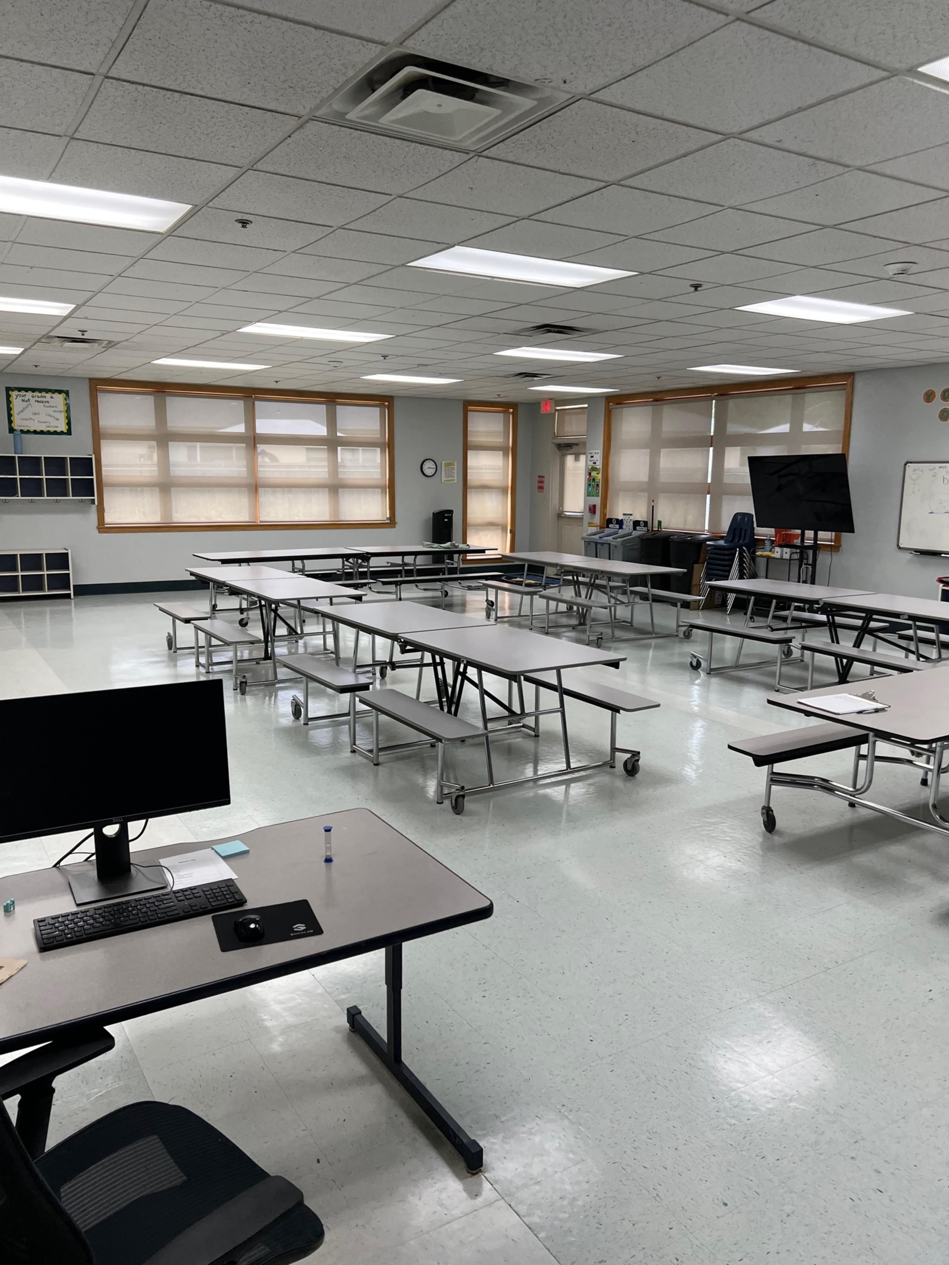 An empty classroom with several tables arranged in rows, a computer on a desk, and large windows allowing natural light.