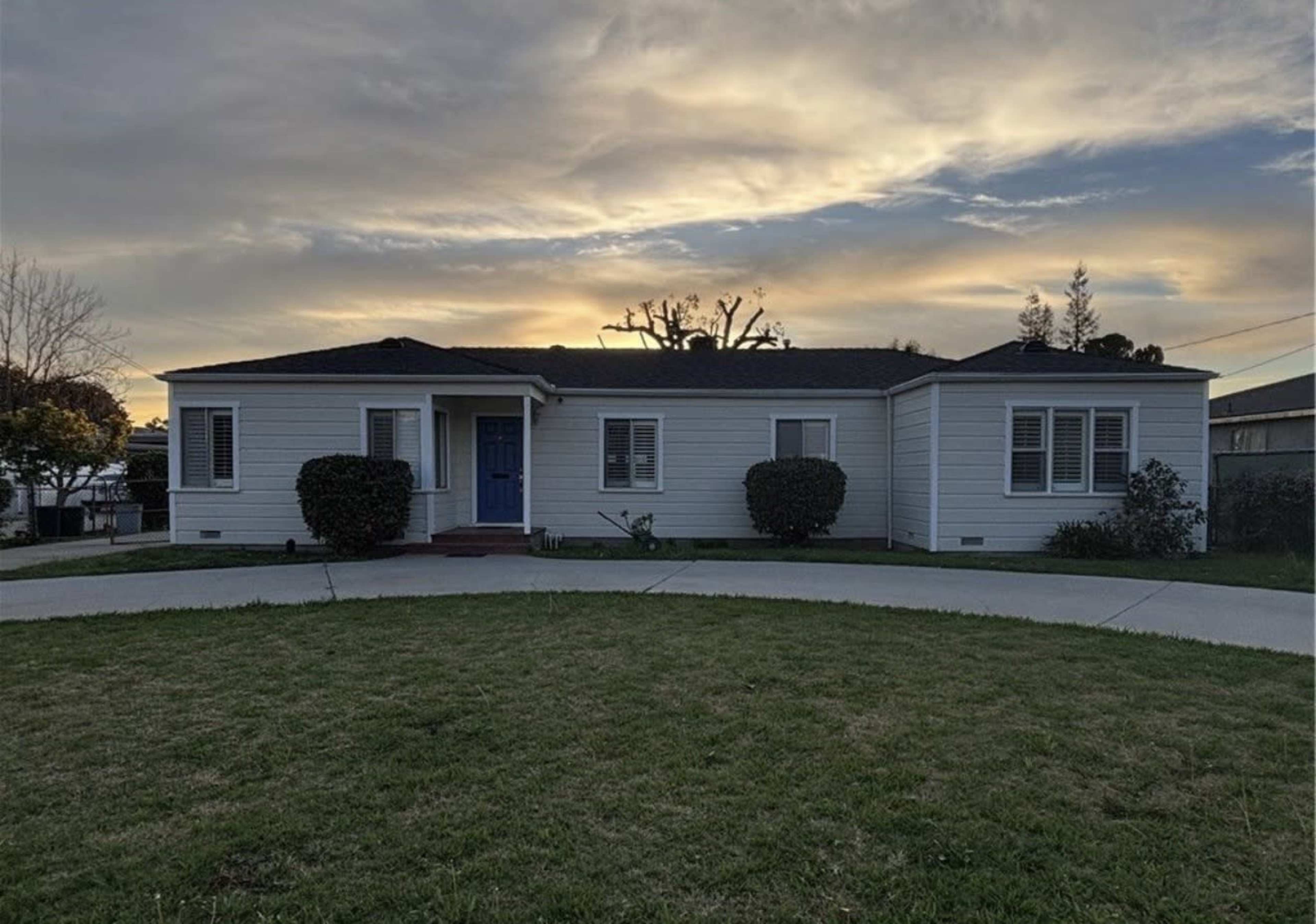 A single-story white house with a blue front door is situated on a grassy lawn under a cloudy sunset sky.