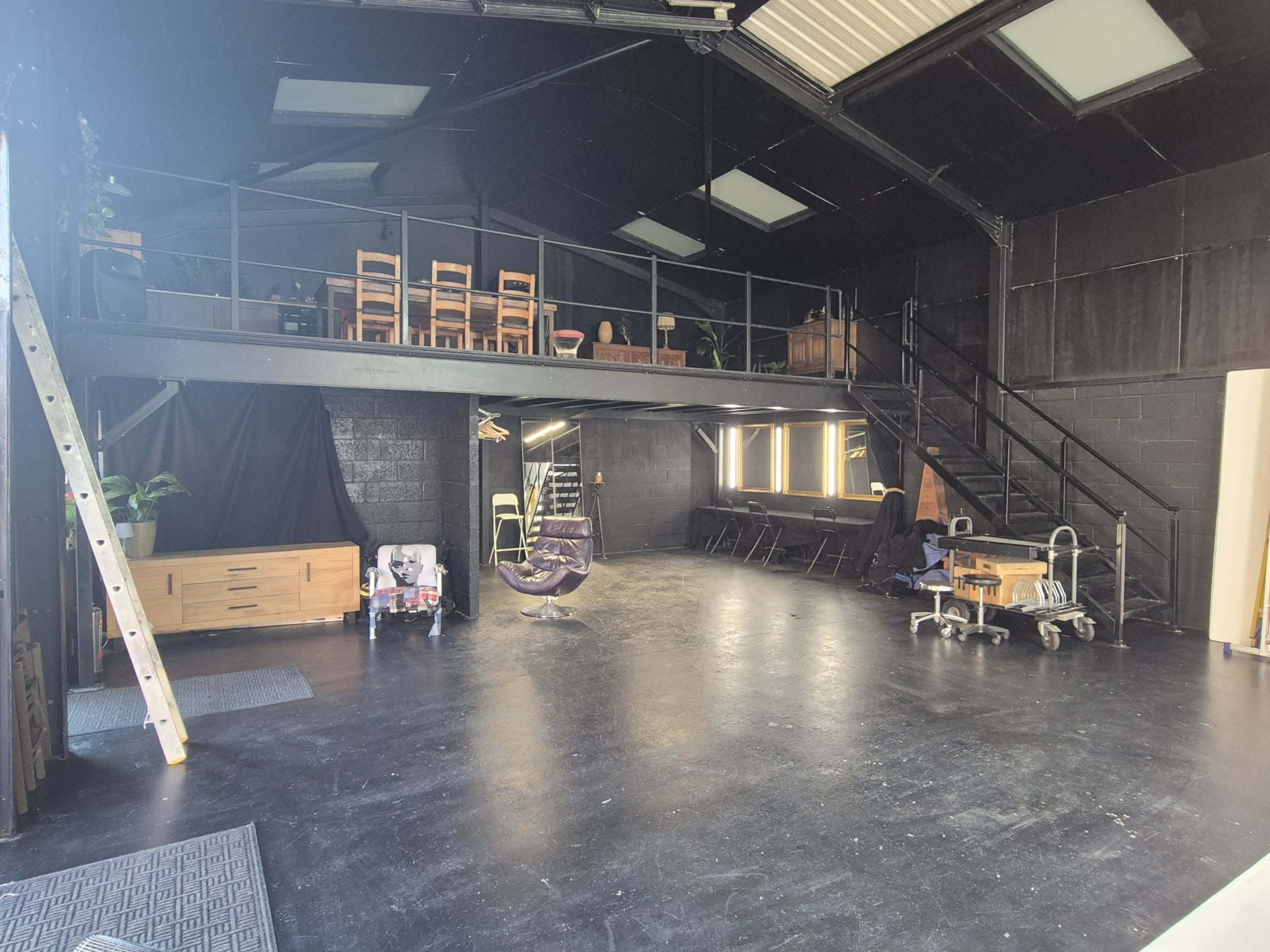 The image shows an empty studio space with high ceilings, a mezzanine area with chairs, and various furnishings on the ground floor.