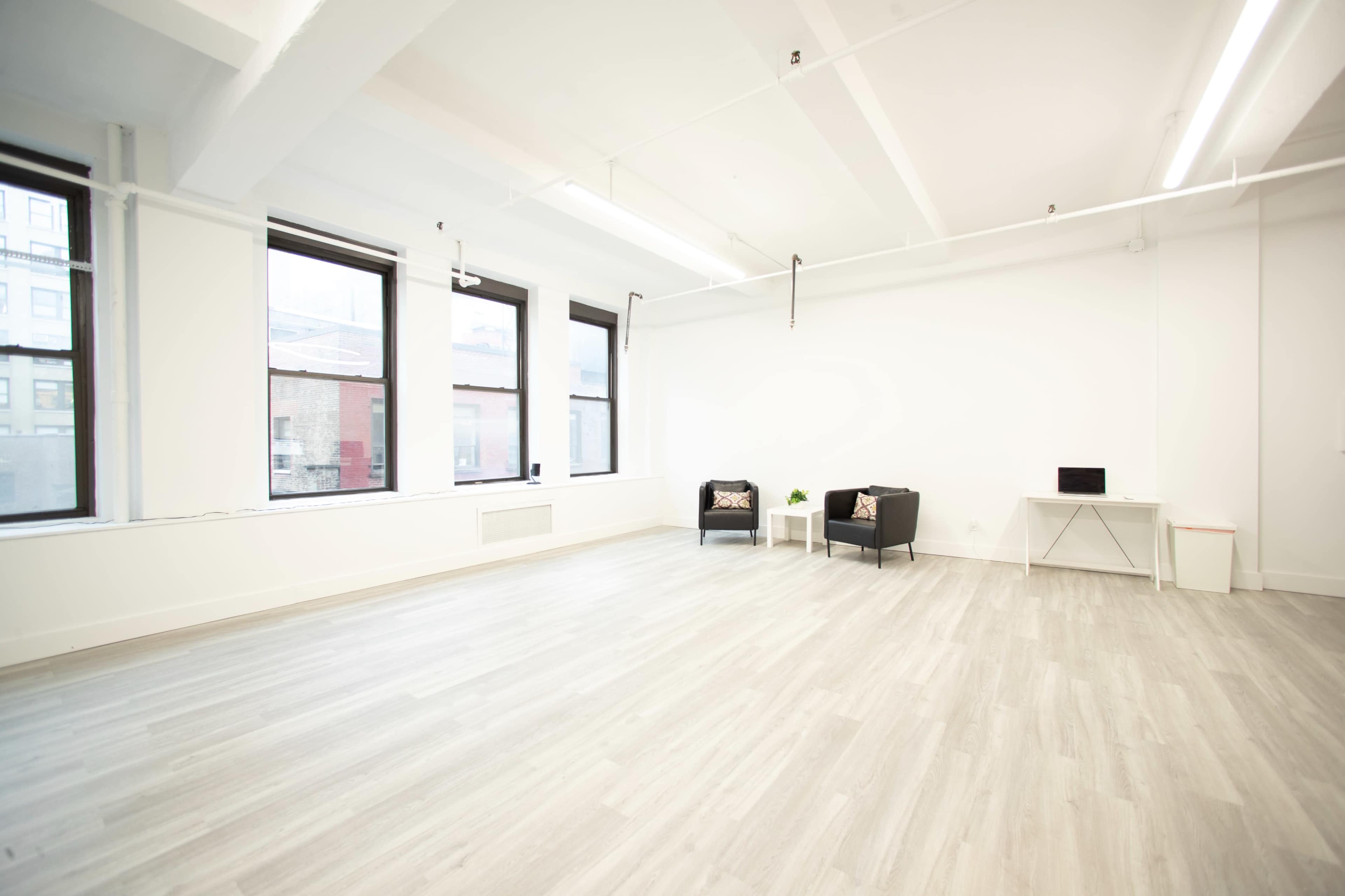 The image shows a spacious, minimally furnished room with two black chairs, a small table, and a desk against a white wall, illuminated by natural light from large windows.