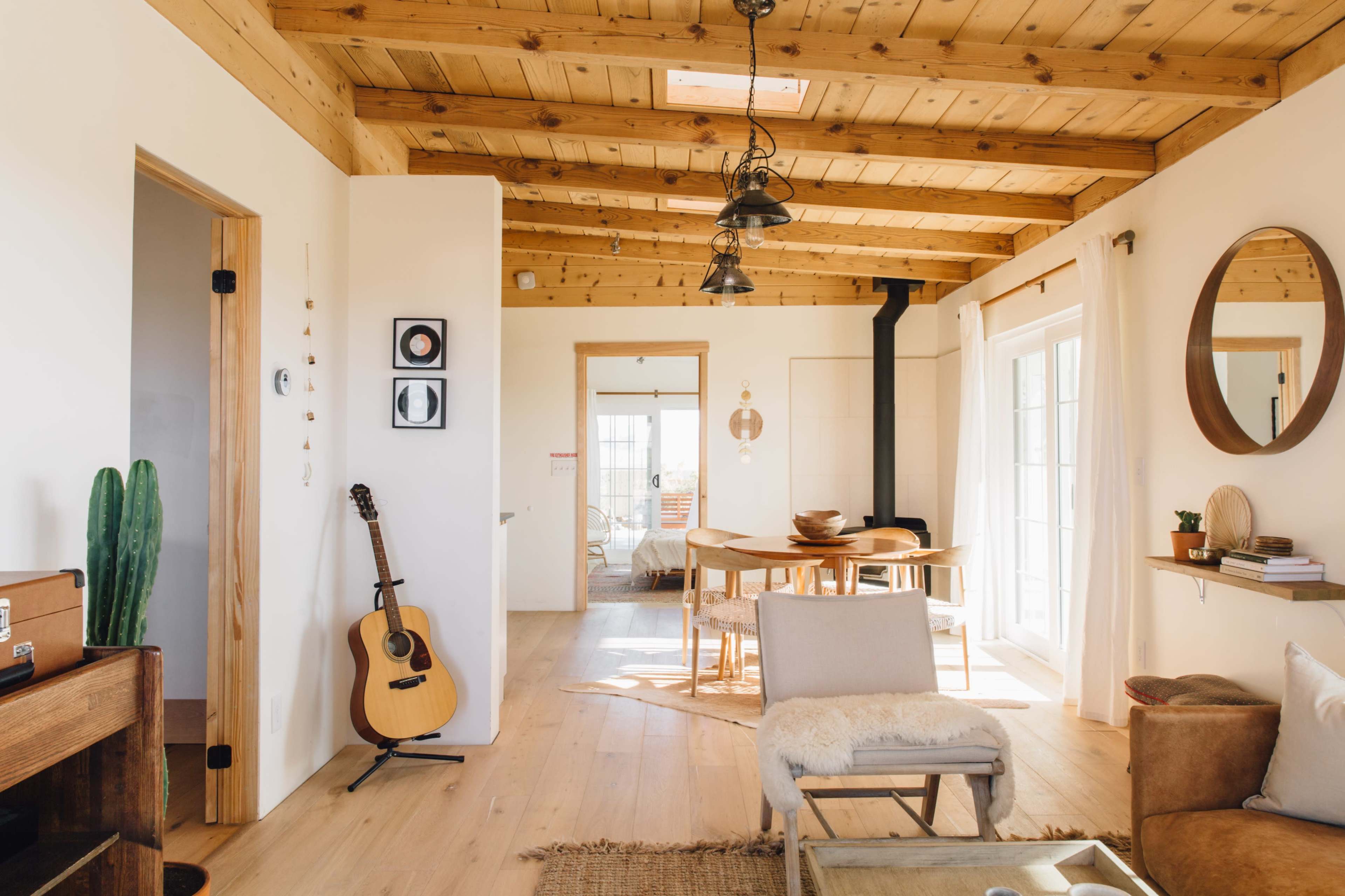 A bright, open living space with wooden beams, a guitar leaning against the wall, and a round dining table surrounded by chairs.