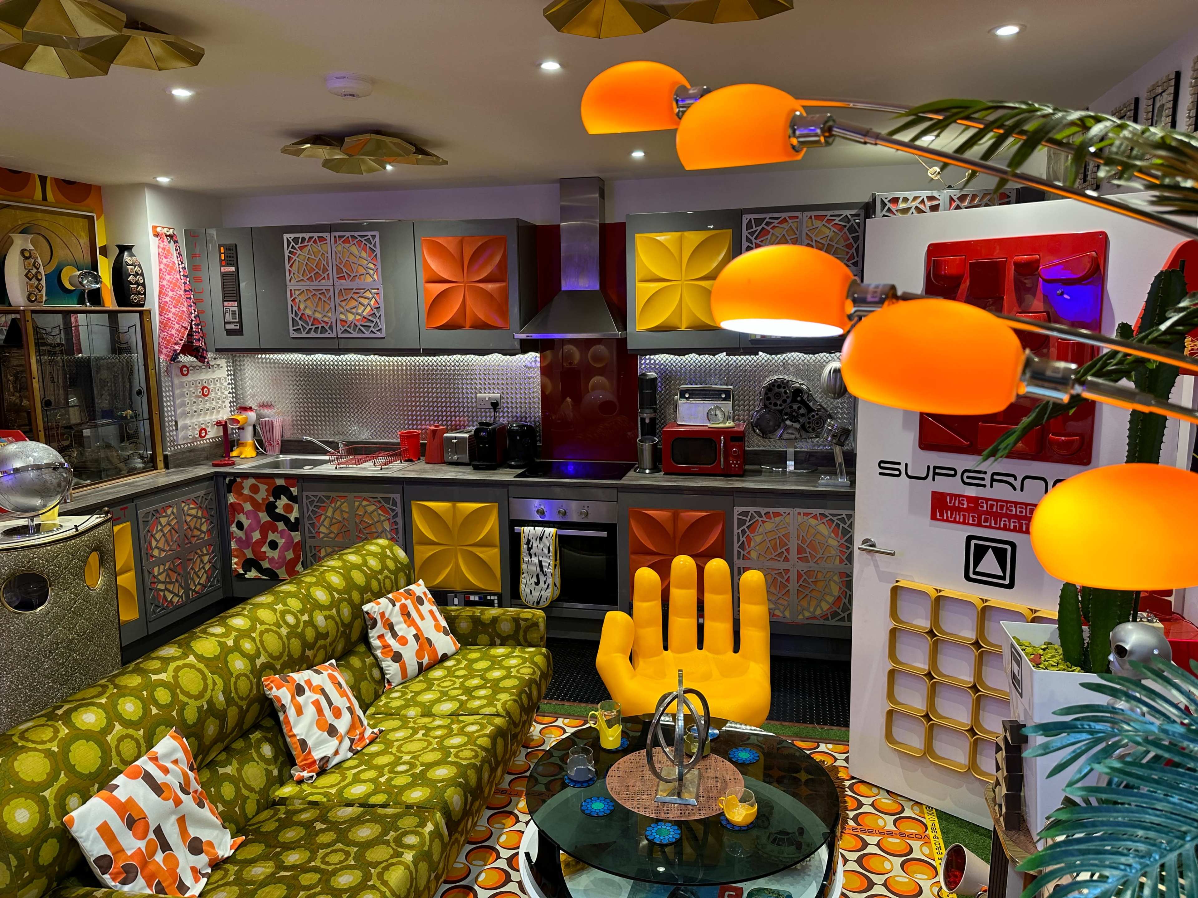 A colorful retro-styled kitchen and living area featuring a patterned sofa, large orange lamps, and decorative wall panels.