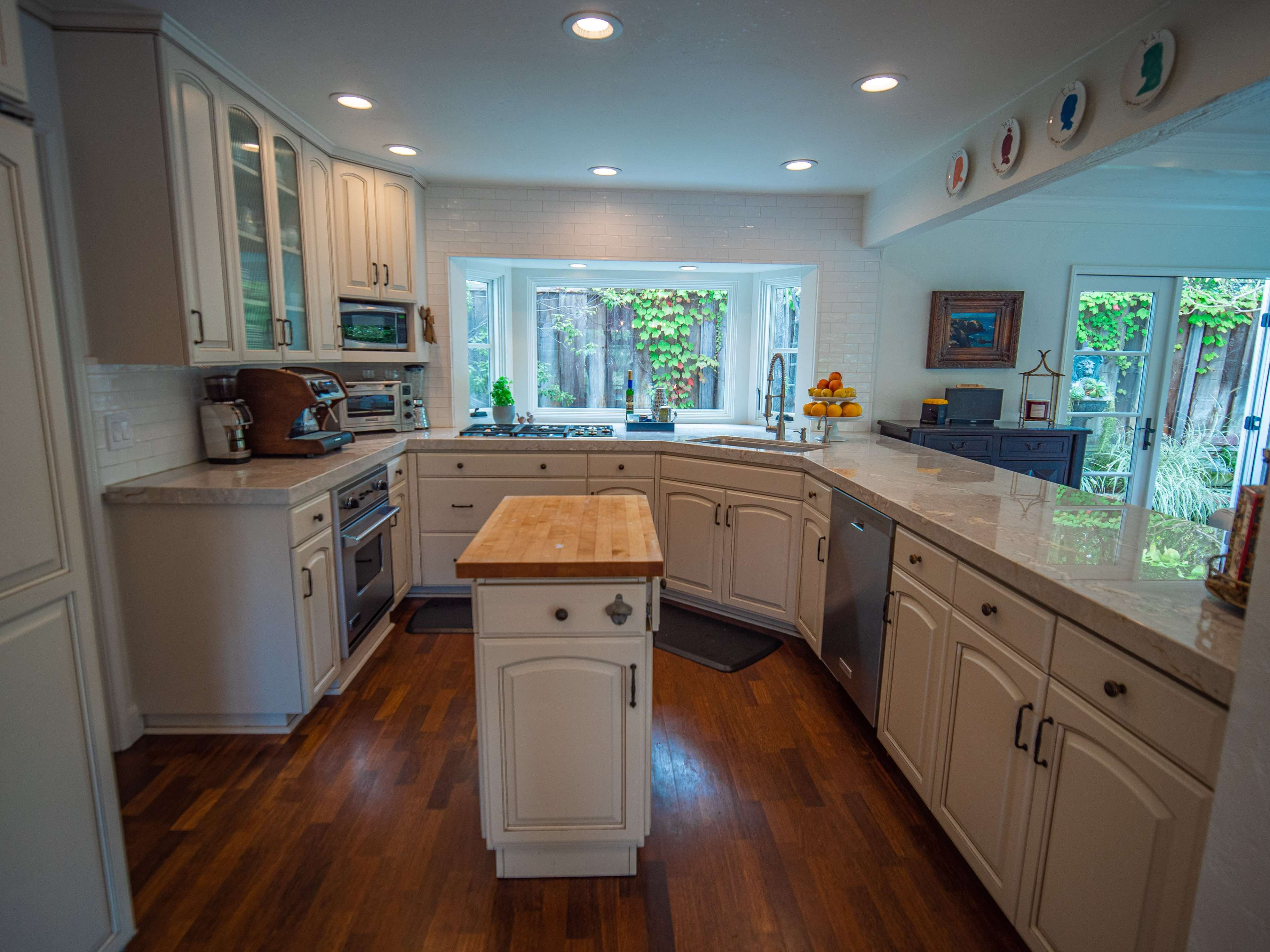 A modern kitchen with white cabinetry, a central wooden island, and a large window overlooking a garden.