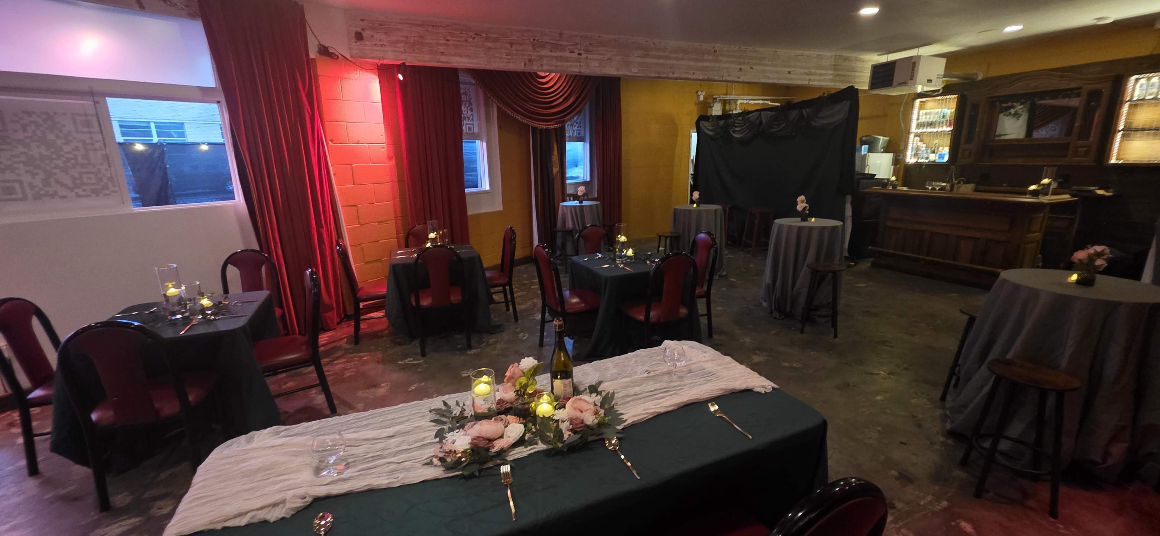 The image shows a dimly lit dining area with round tables covered in gray cloths, arranged around a central long table adorned with flowers and candles, alongside a bar area in the background.