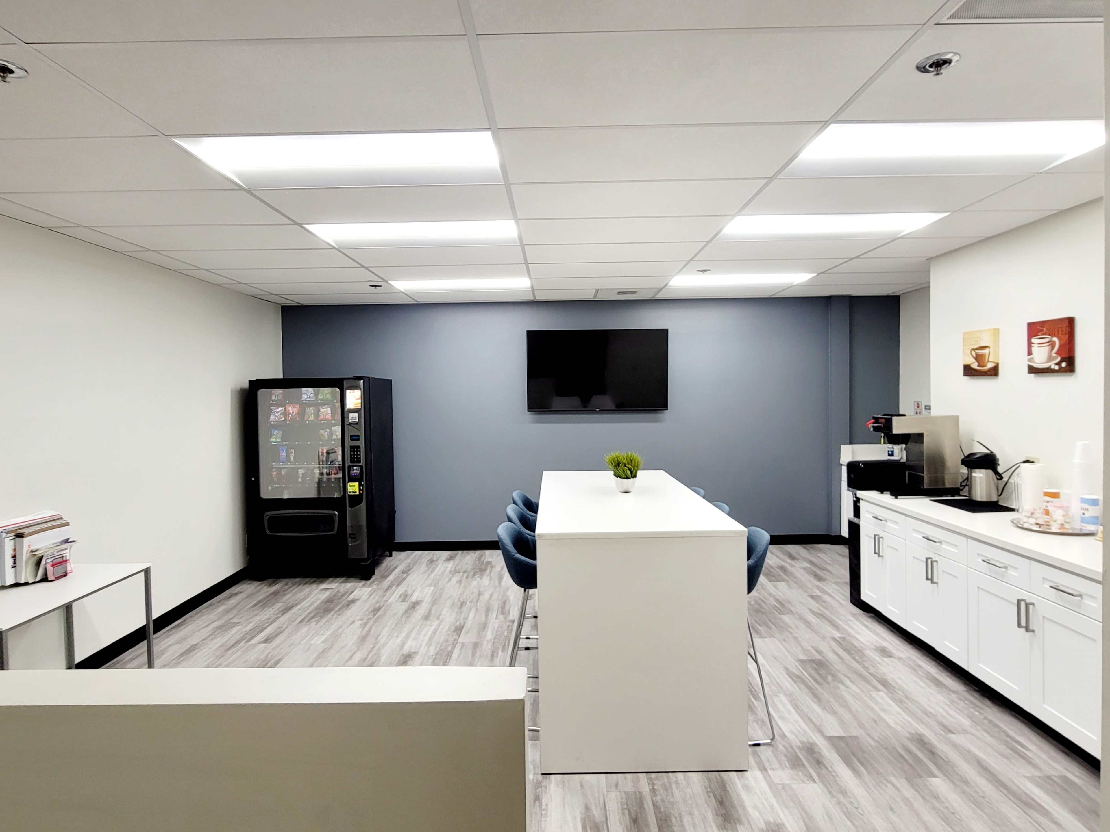 A modern break room with a vending machine, a large table surrounded by chairs, and a coffee station along one wall, all under bright overhead lighting.