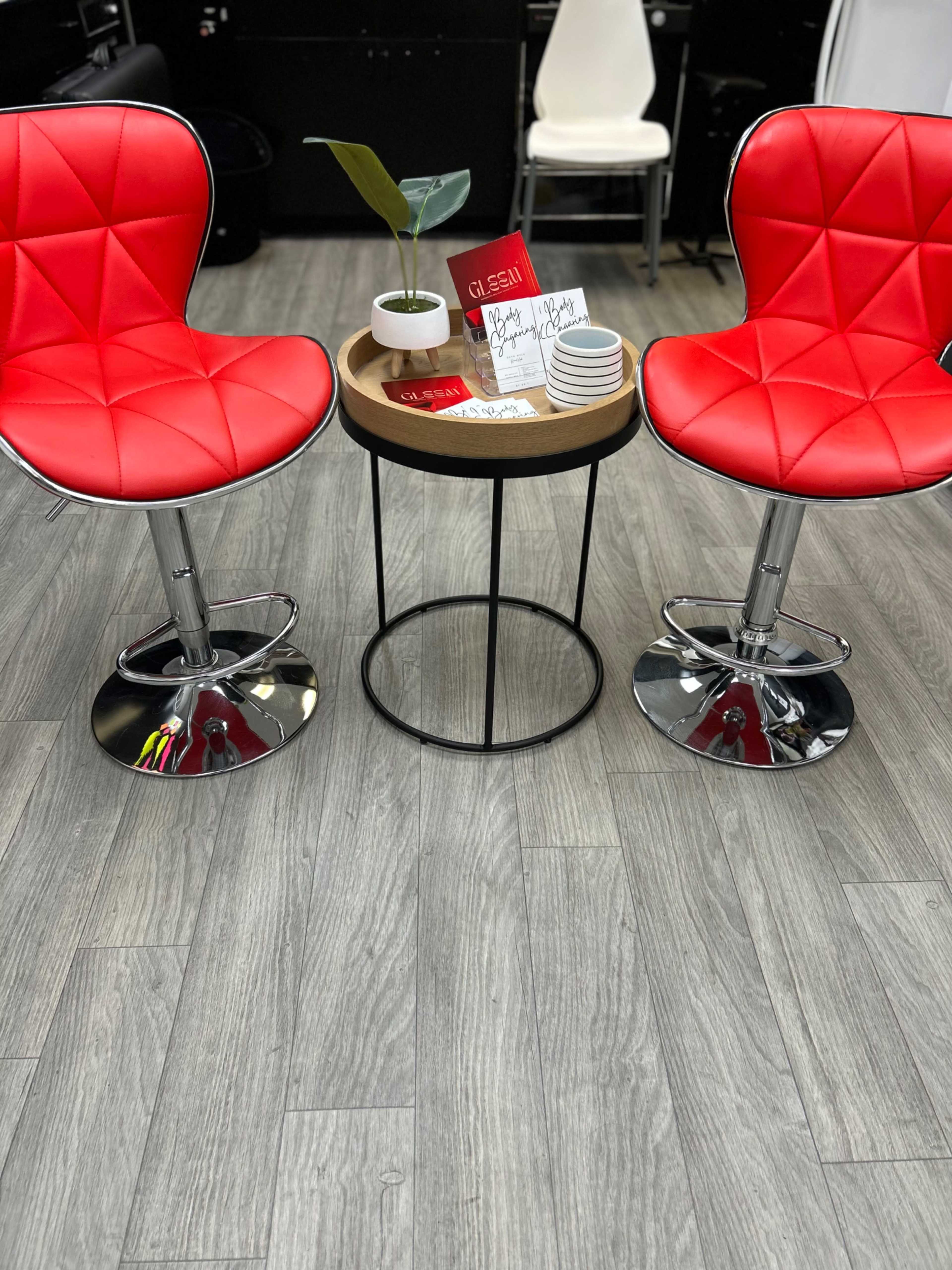 Two red swivel chairs flank a small round table with a potted plant and stationery on a wood-patterned floor.