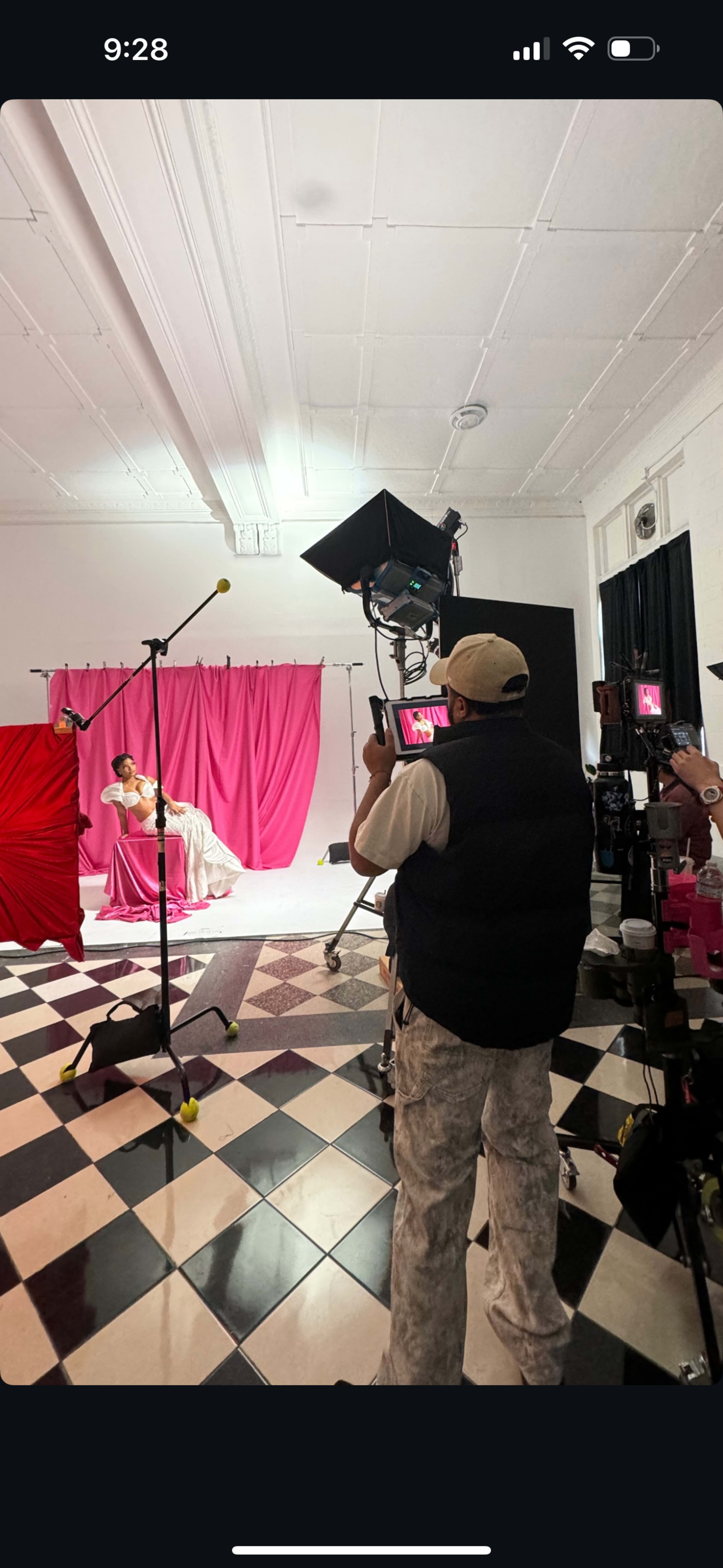 A photographer captures a model posing in a studio with pink backdrops and black-and-white checkered flooring.