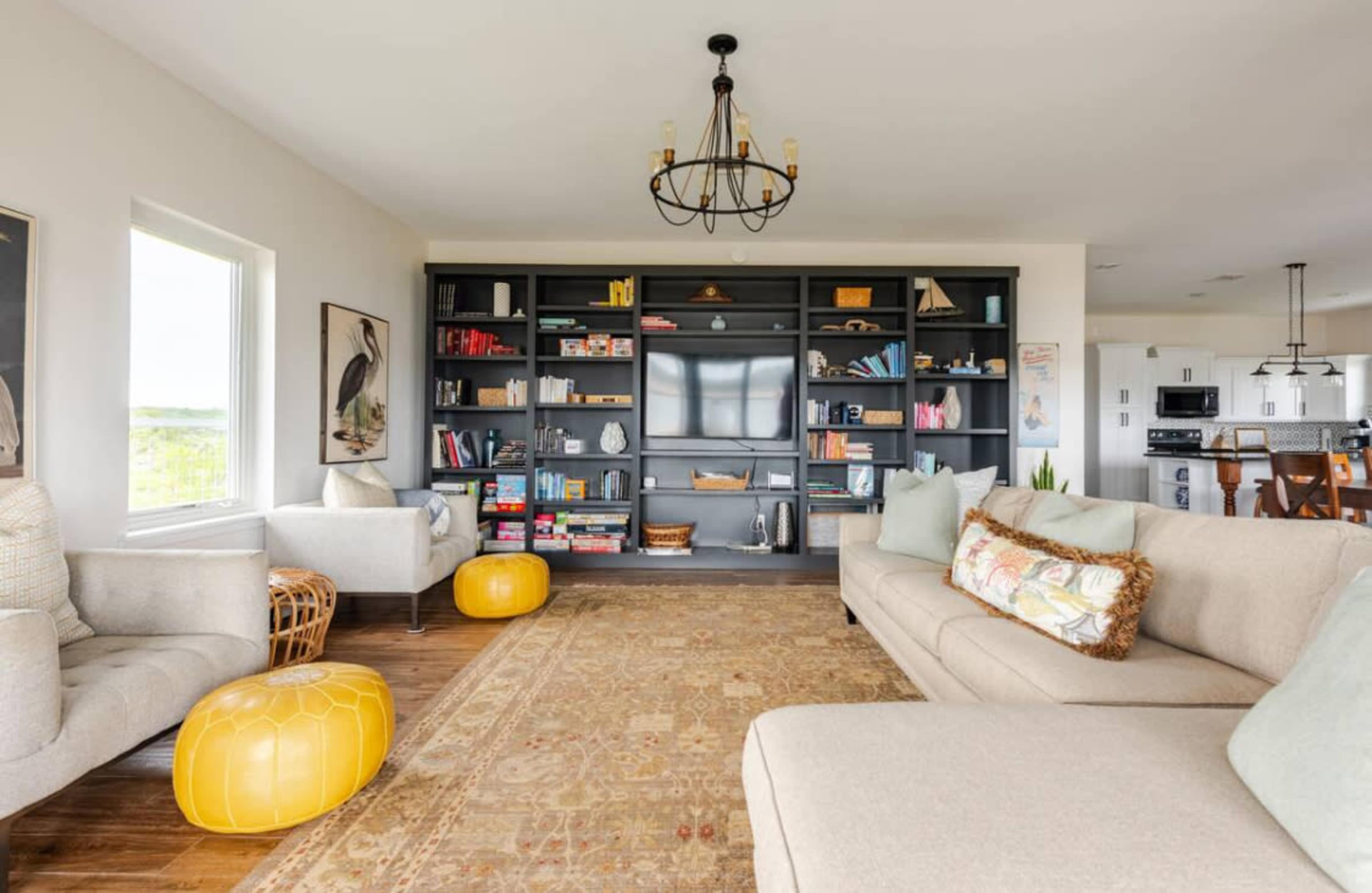 A spacious living room features a light-colored sofa, two armchairs, a dark shelving unit filled with books and decorative items, and yellow poufs on a patterned area rug.