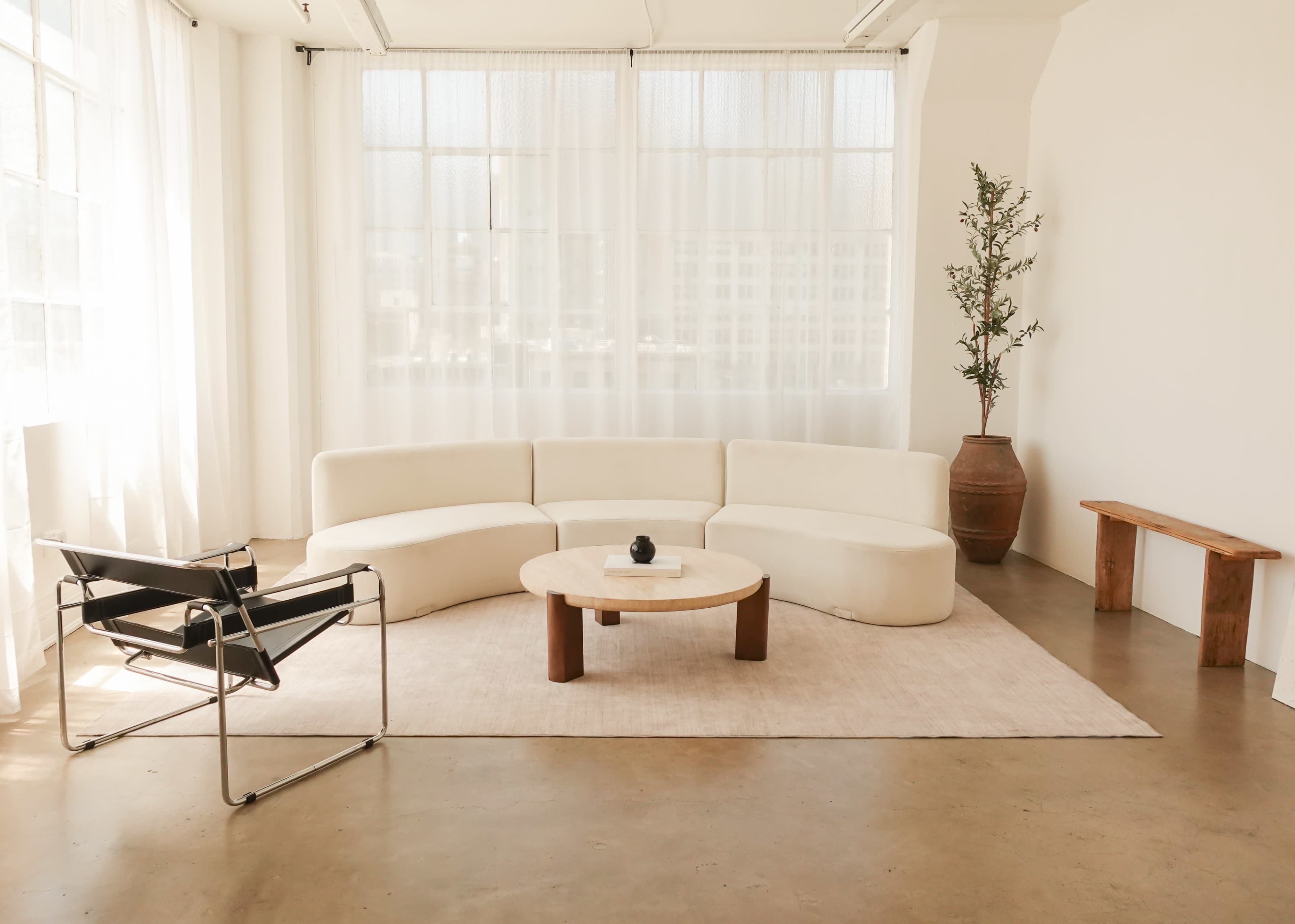 The image shows a modern, minimalist living space featuring a curved white sofa, a round coffee table, a black chair, and a potted plant near a large window with sheer curtains.