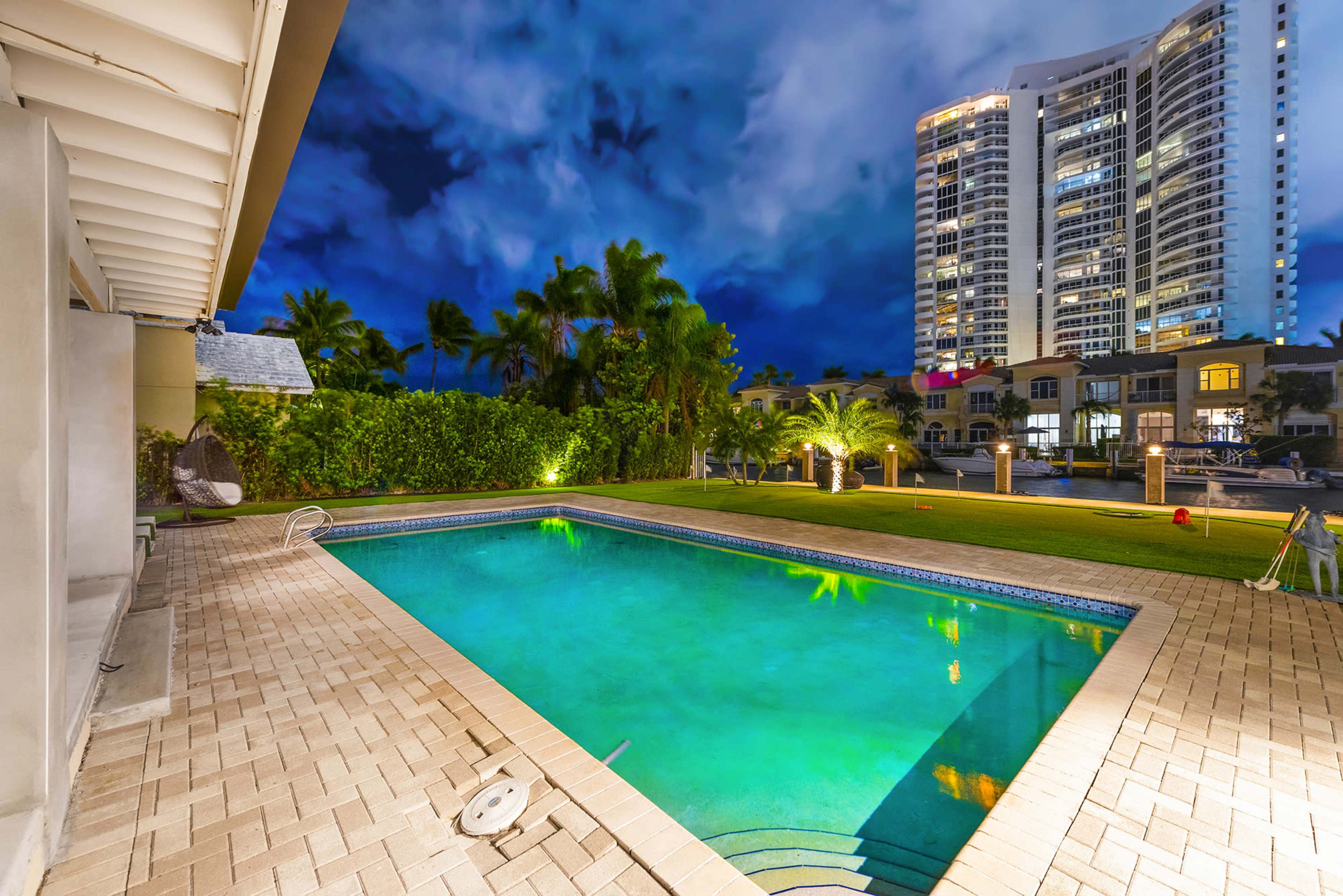 A well-lit swimming pool surrounded by palm trees and modern buildings under a dramatic night sky.