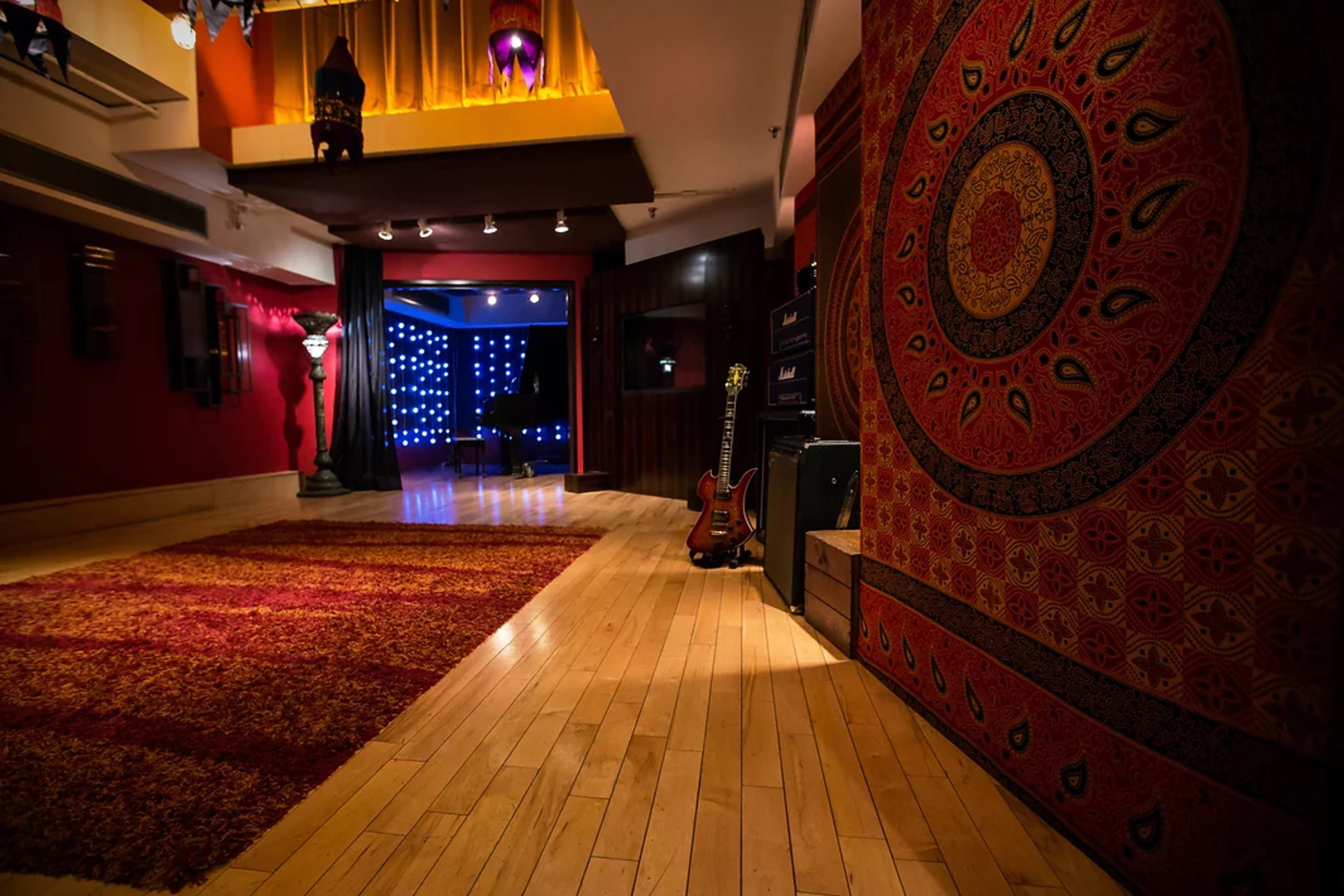 The image shows a spacious room with wooden flooring, a large red rug, decorative wall patterns, and a guitar leaning against an amplifier.