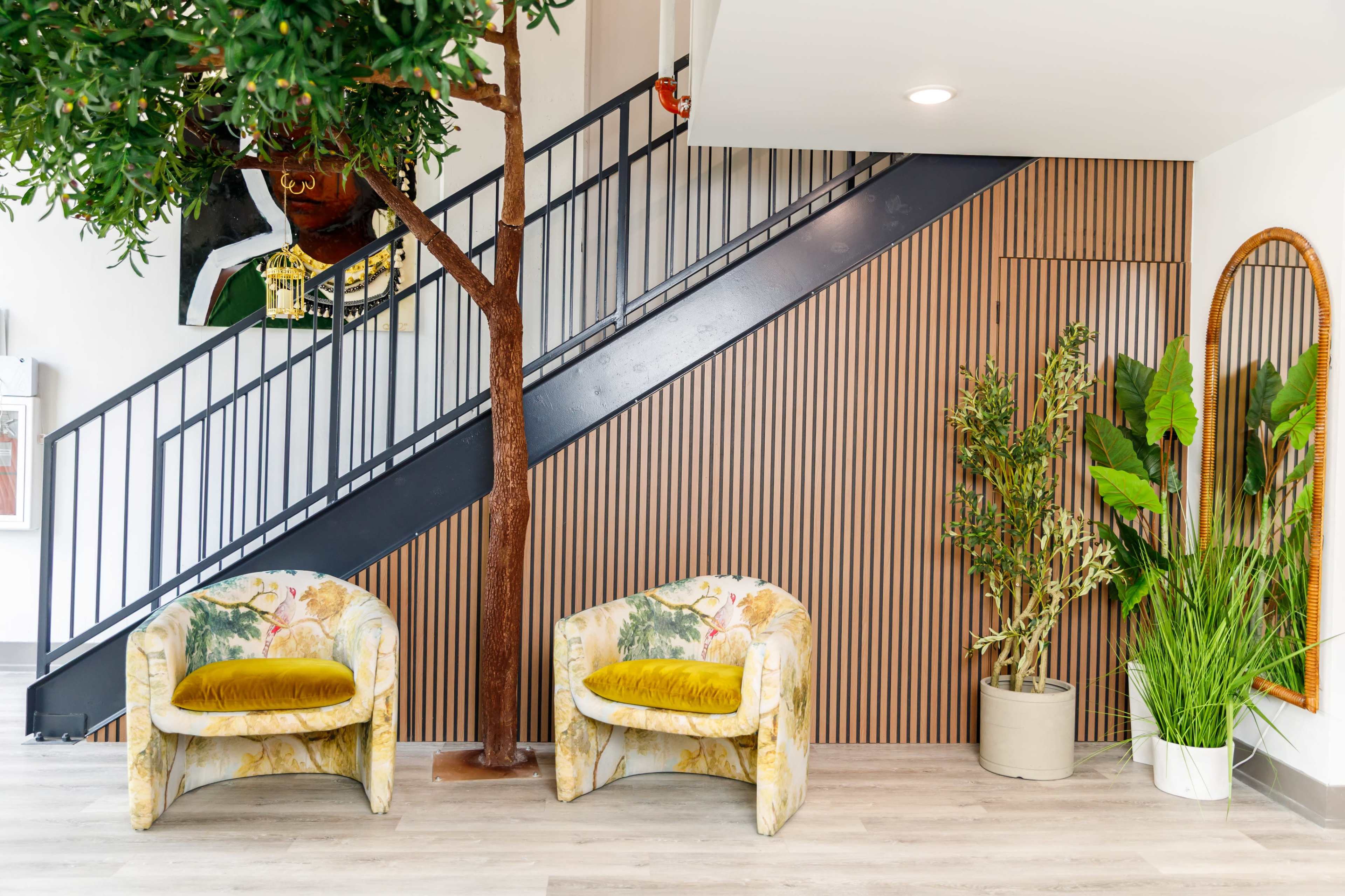 The image shows a modern interior with two patterned chairs, a wooden staircase, and plants located beside a textured wooden wall.