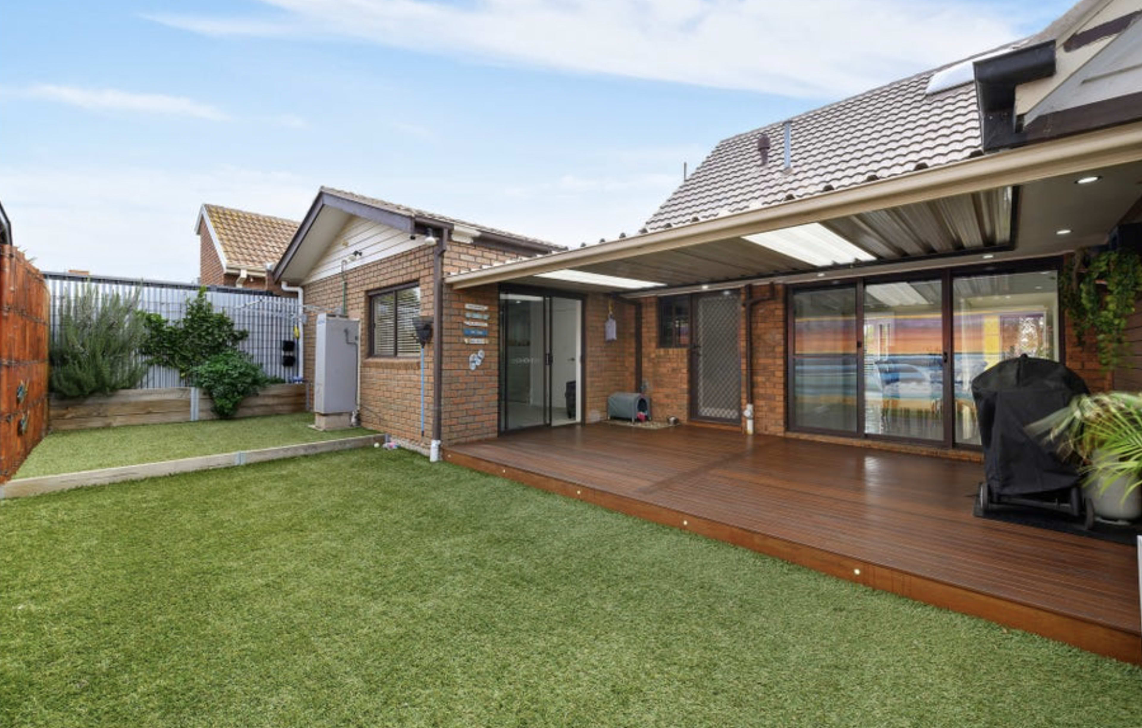 The image shows a well-maintained backyard featuring a wooden deck, artificial grass, and a brick house with large glass doors leading to the outdoor space.