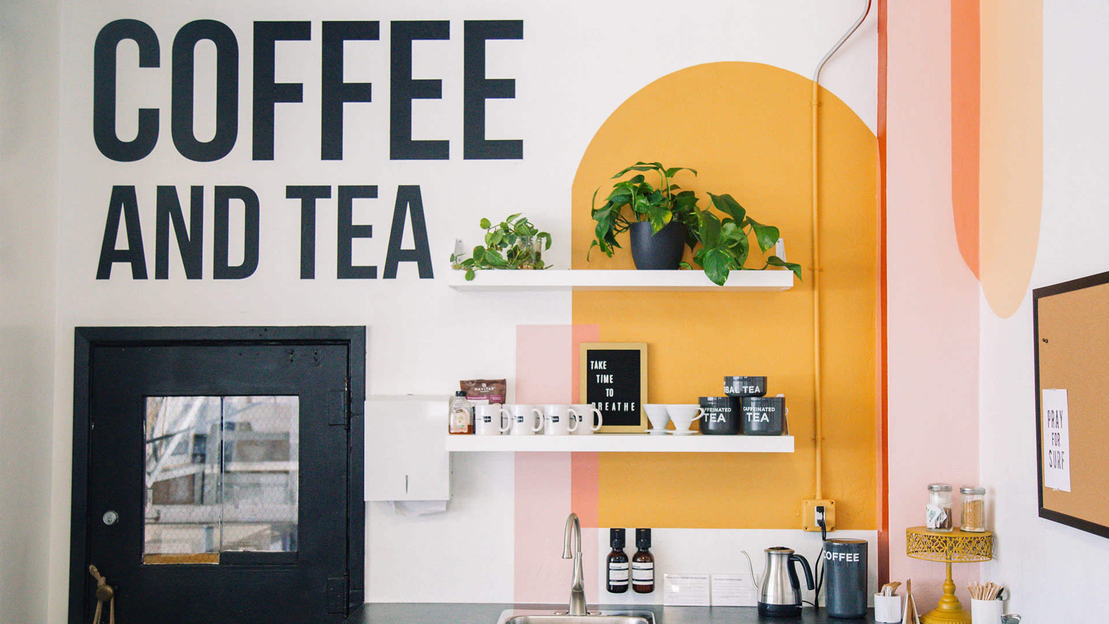 A café interior with a wall featuring bold text that reads "COFFEE AND TEA," alongside shelves displaying plants and various mugs.
