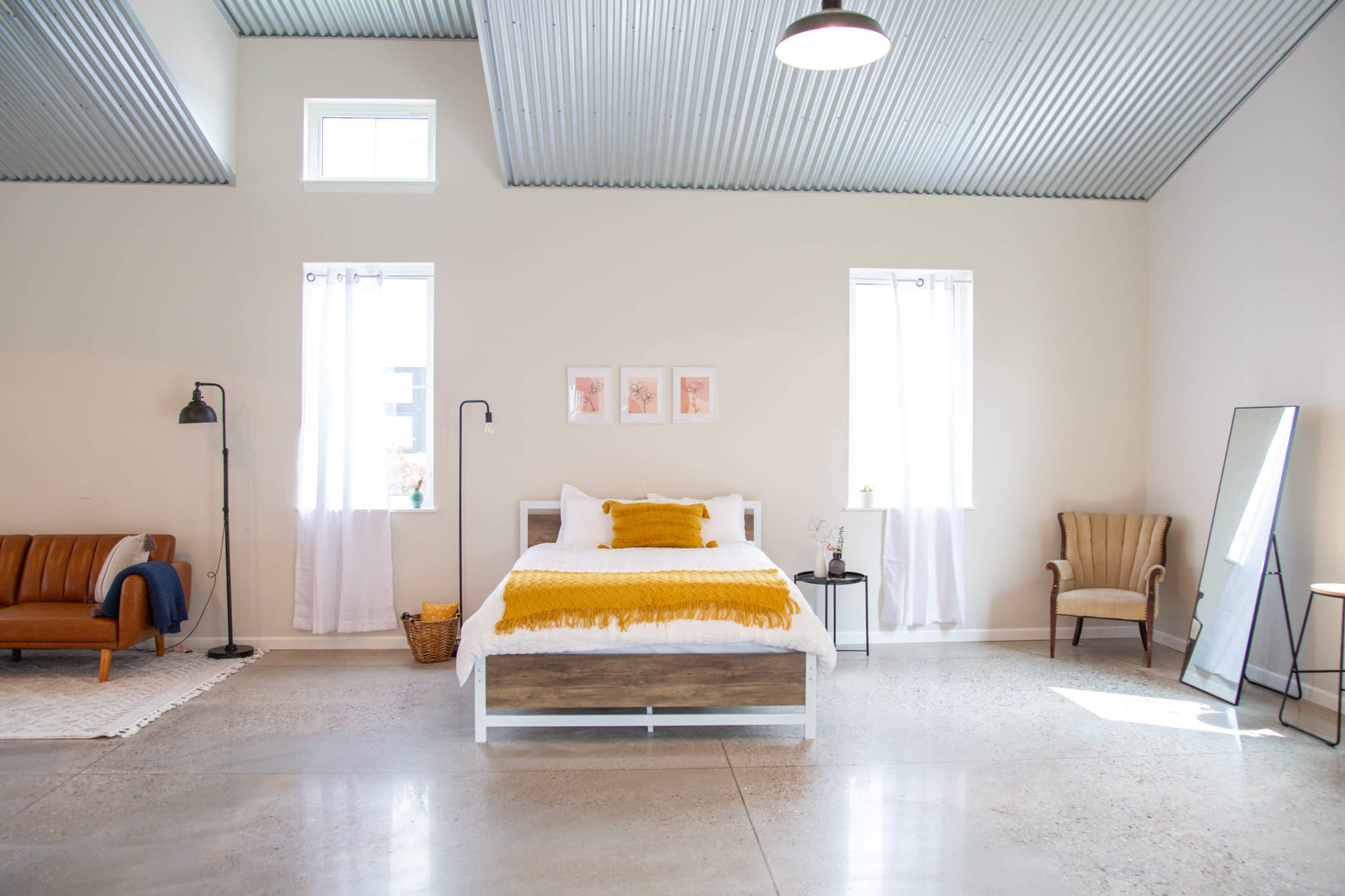 A spacious room features a bed with a yellow throw, accompanied by a seating area and large windows with white curtains.