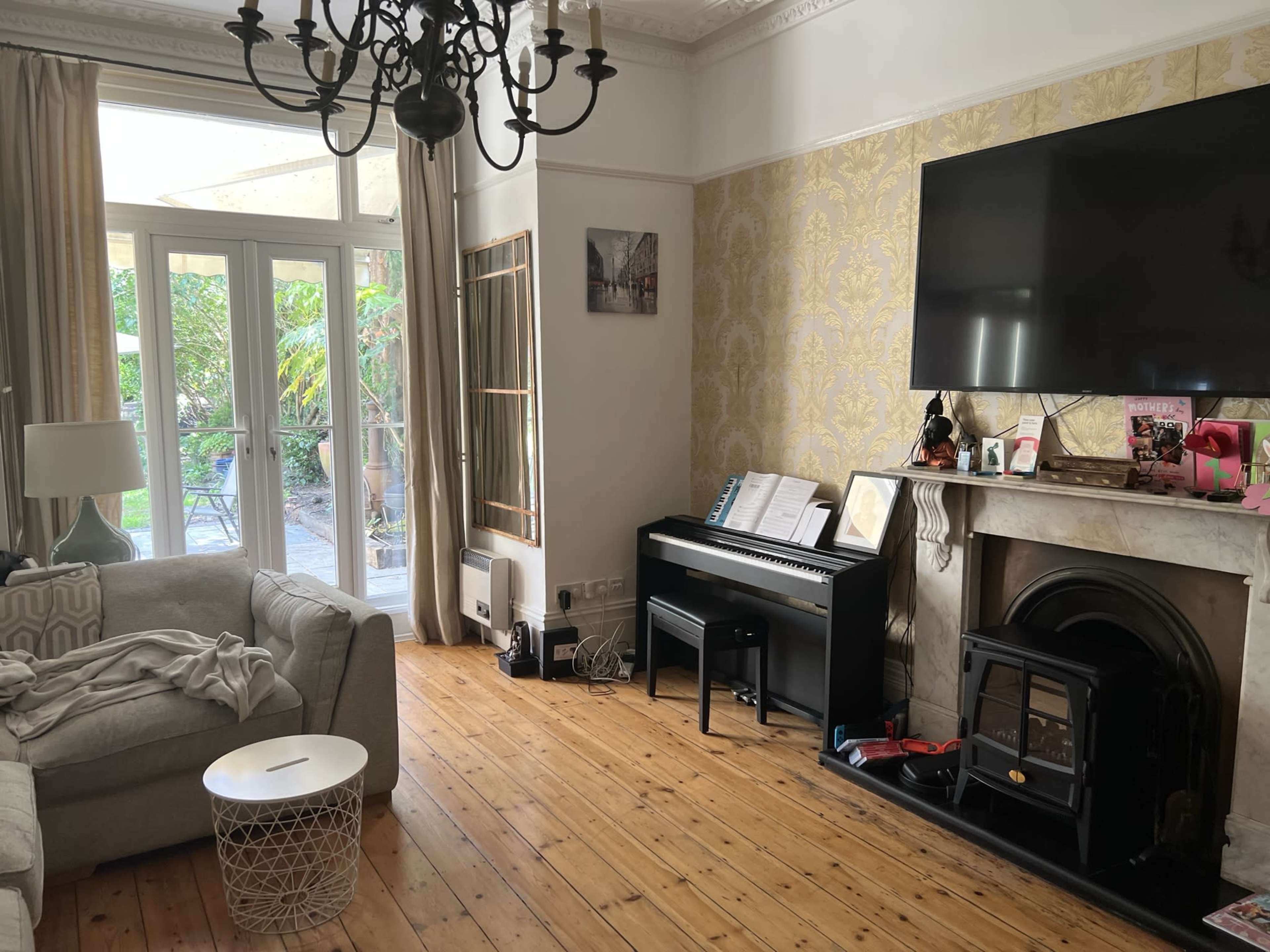 The living room features a chandelier, a large television mounted on the wall, a piano, and a cozy sofa with a throw on its arm, along with a view of a garden through glass doors.