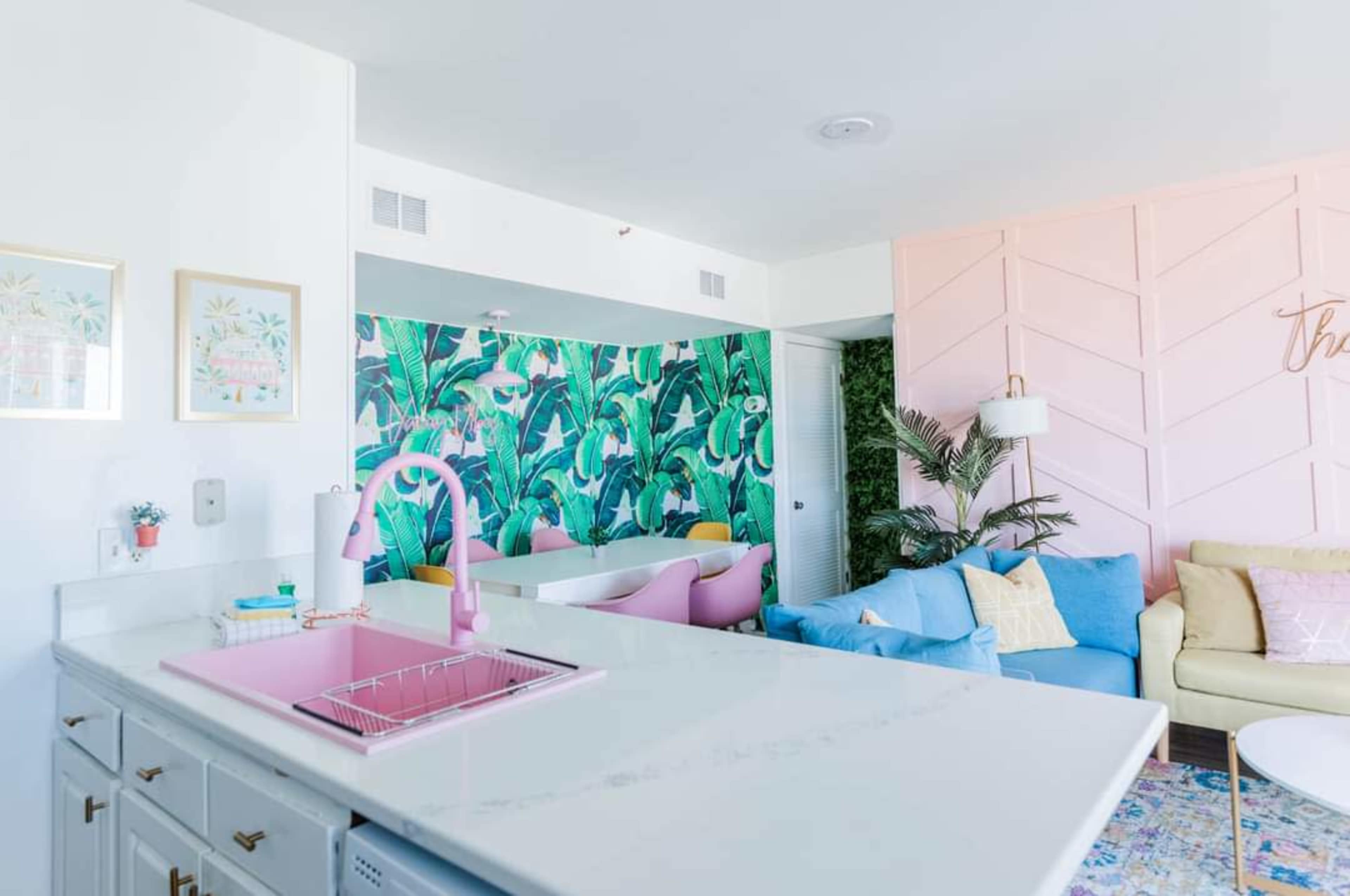 A modern kitchen with a pink faucet and a marble countertop, leading to a vibrant living area adorned with a tropical wallpaper and colorful furniture.