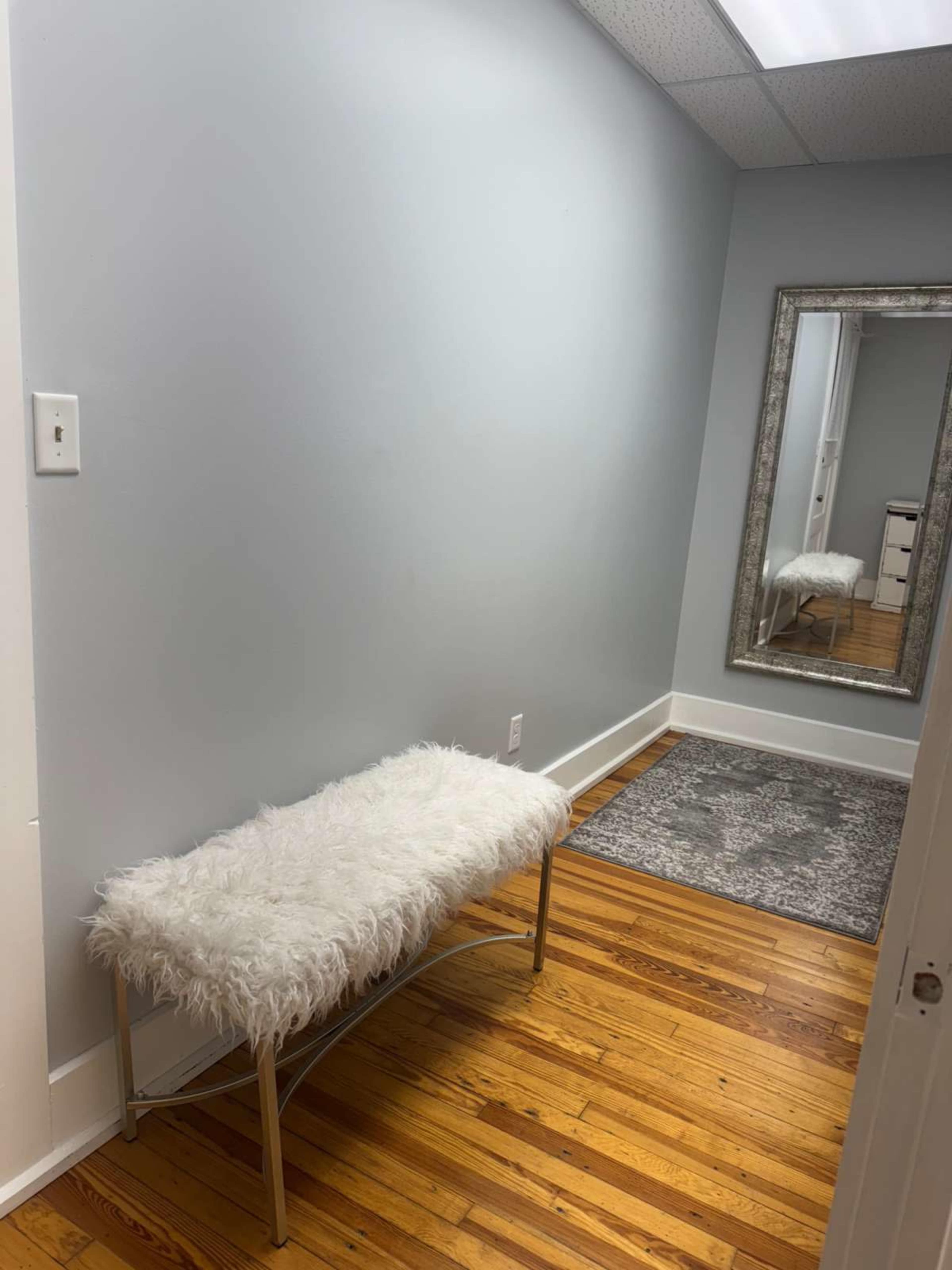 The image shows a narrow hallway with a white bench, a large mirror, and a gray area rug on a wooden floor.