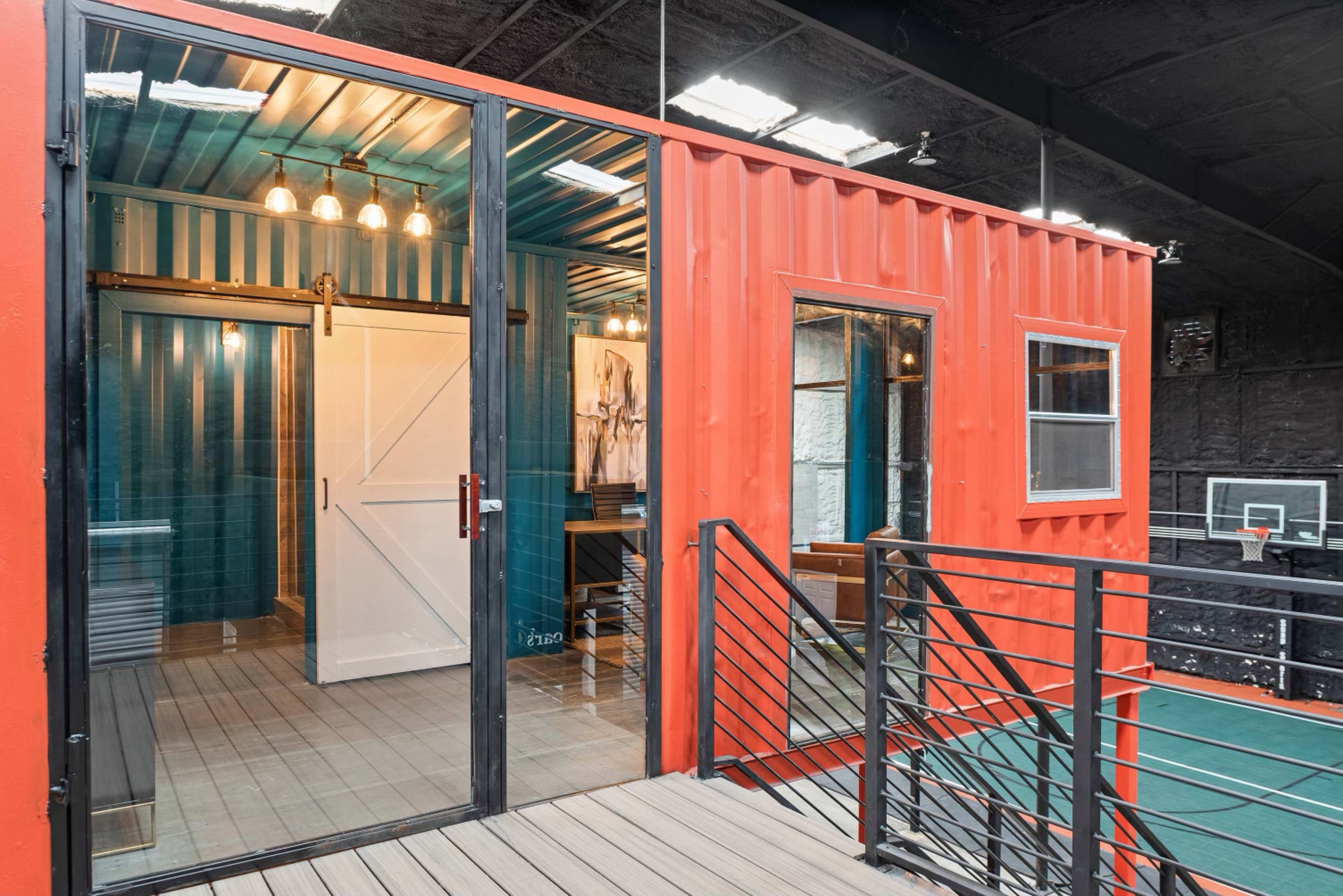 The image shows a modern space constructed from shipping containers, featuring a glass entrance and an outdoor deck, with basketball courts visible in the background.