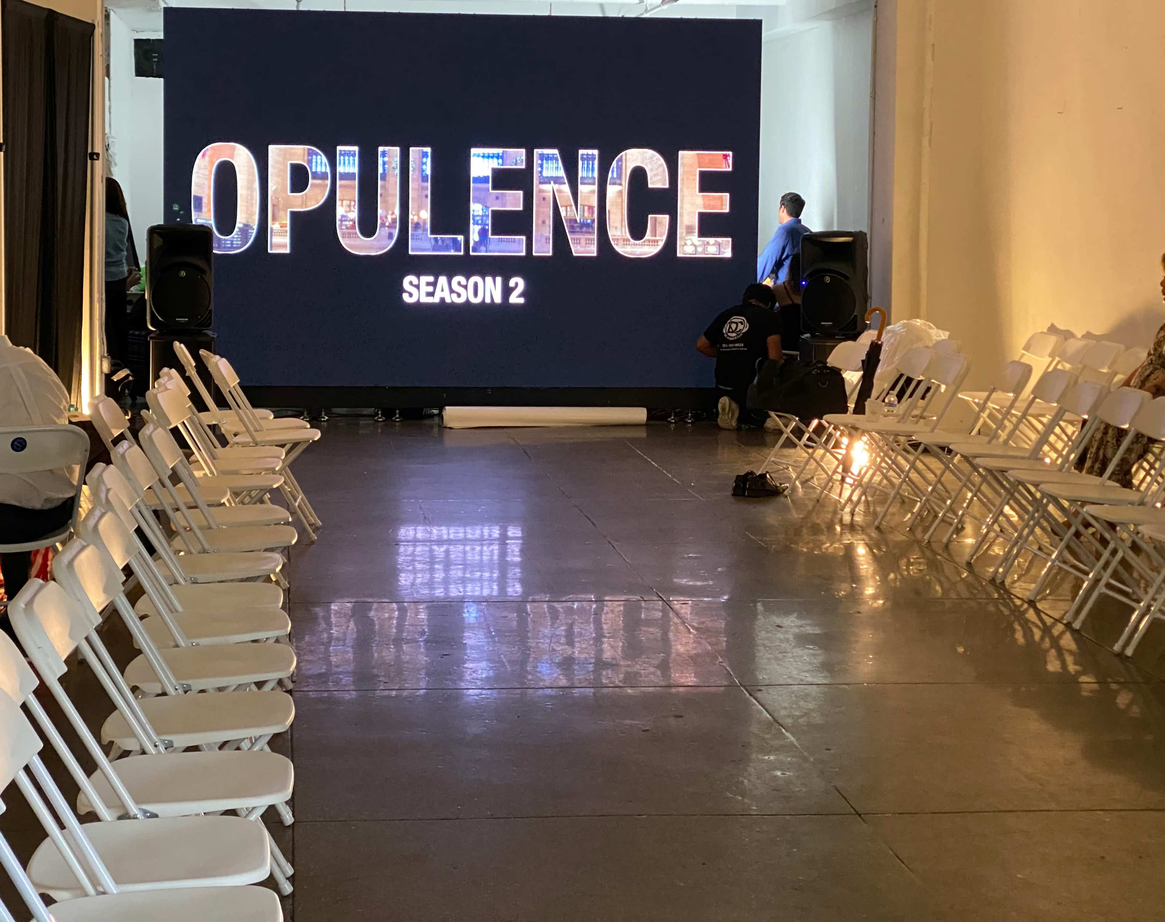 A spacious venue features white folding chairs arranged in two rows facing a large screen displaying "OPULENCE SEASON 2."