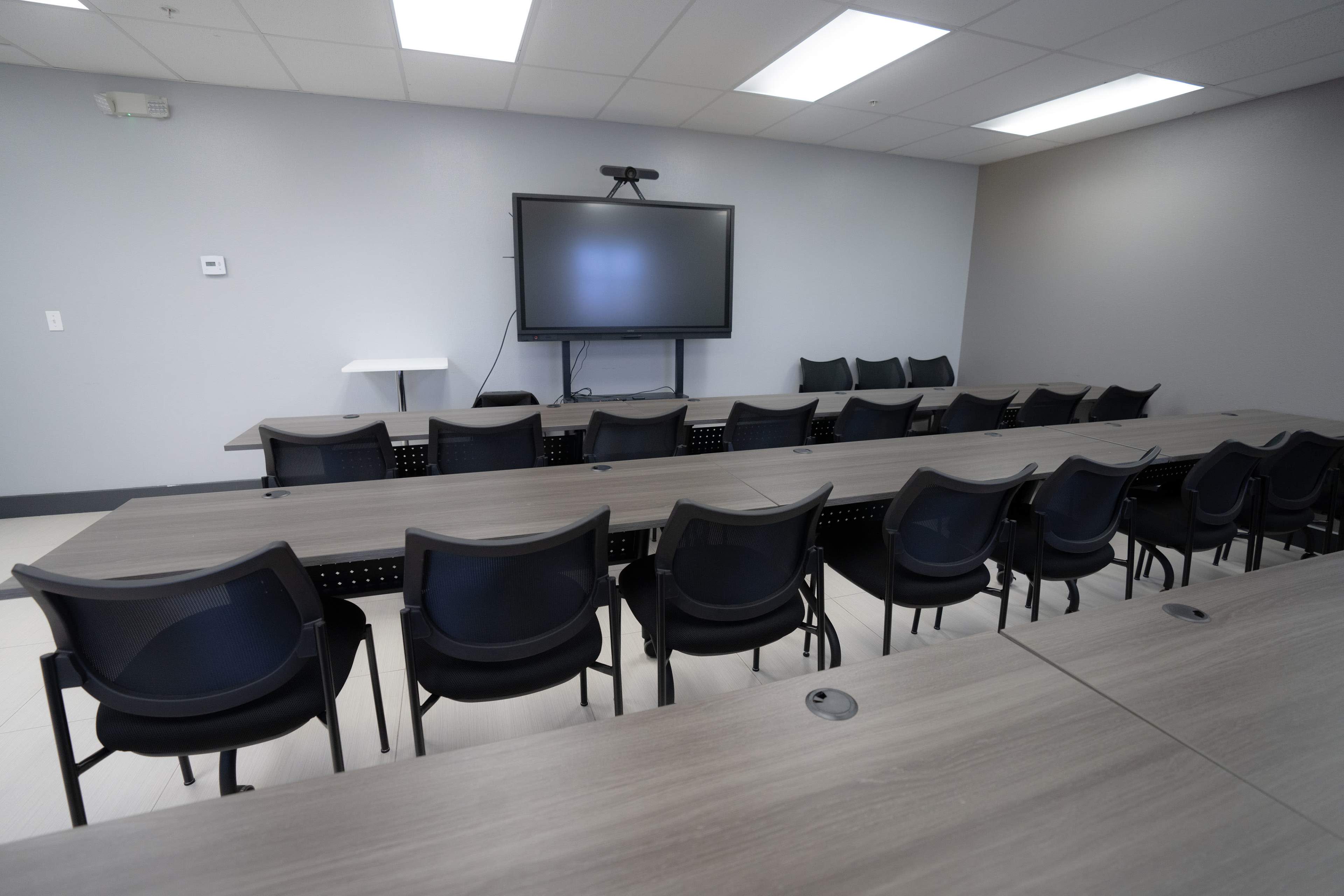 A conference room features several long tables arranged in a U-shape facing a large screen on the wall, with black chairs positioned around the tables.