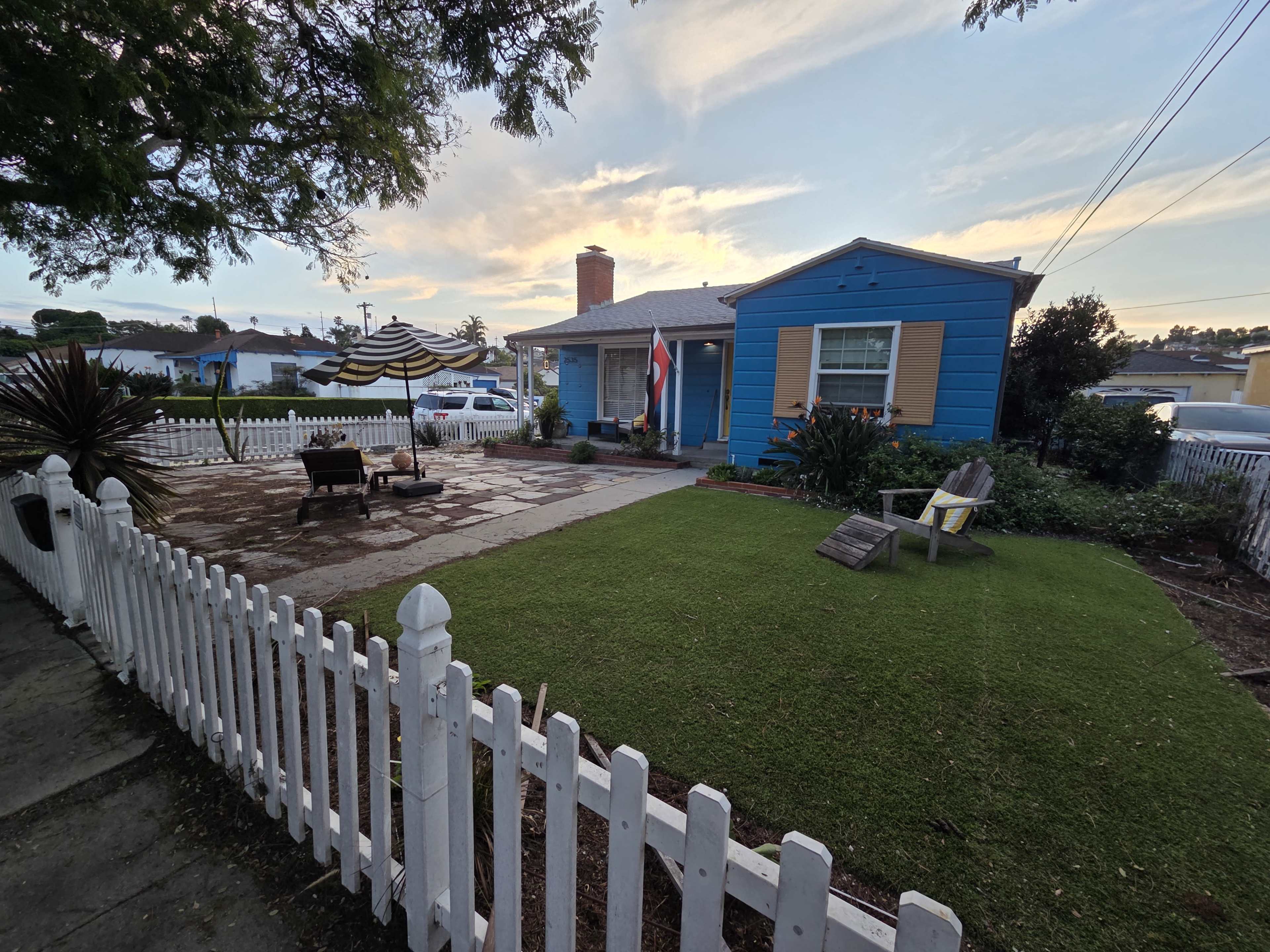 The image shows a blue house with a porch, surrounded by a white picket fence, a landscaped yard, and a stone patio.