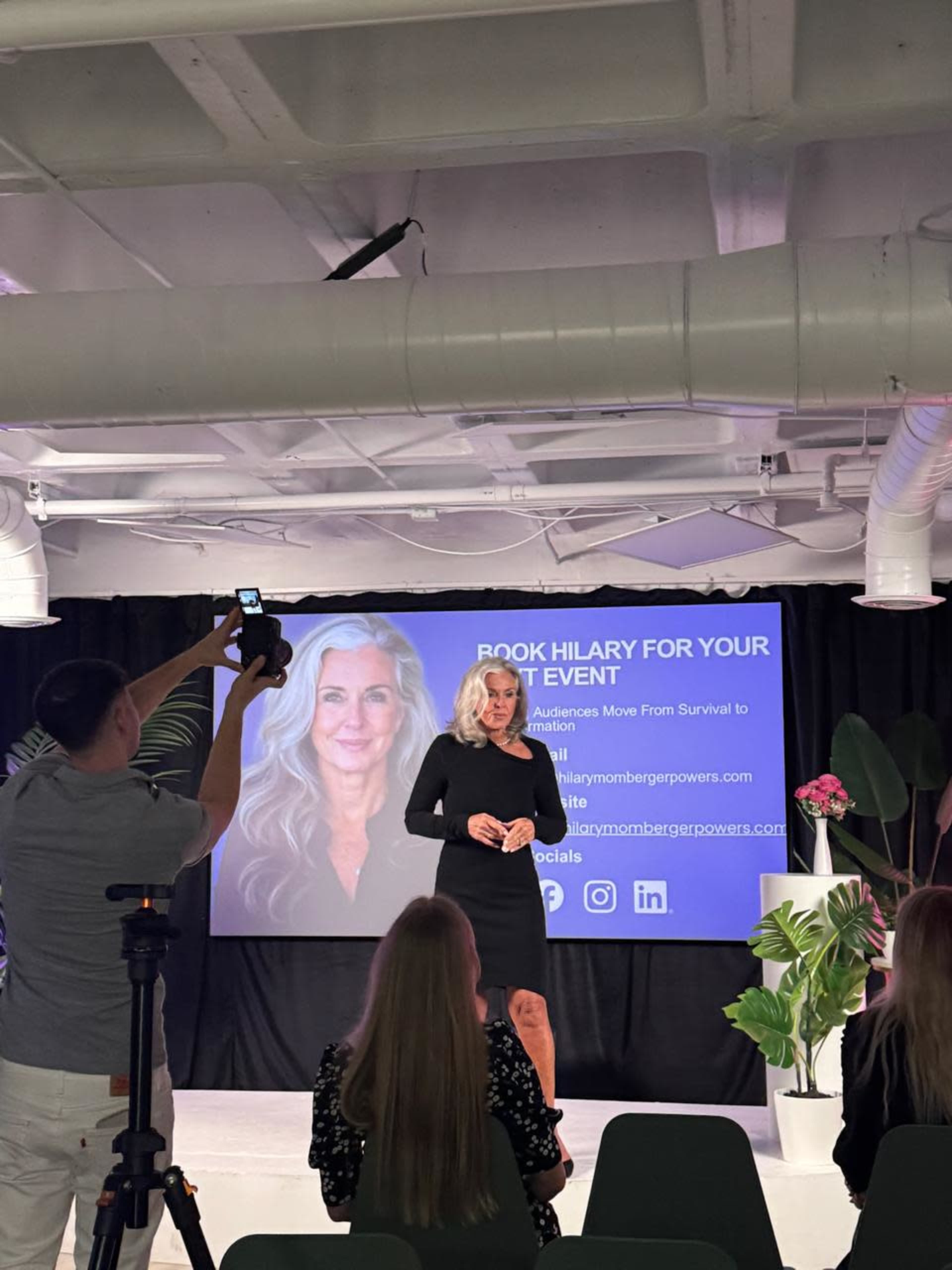 A speaker stands on stage in front of a screen displaying contact details, while an audience member records the presentation.