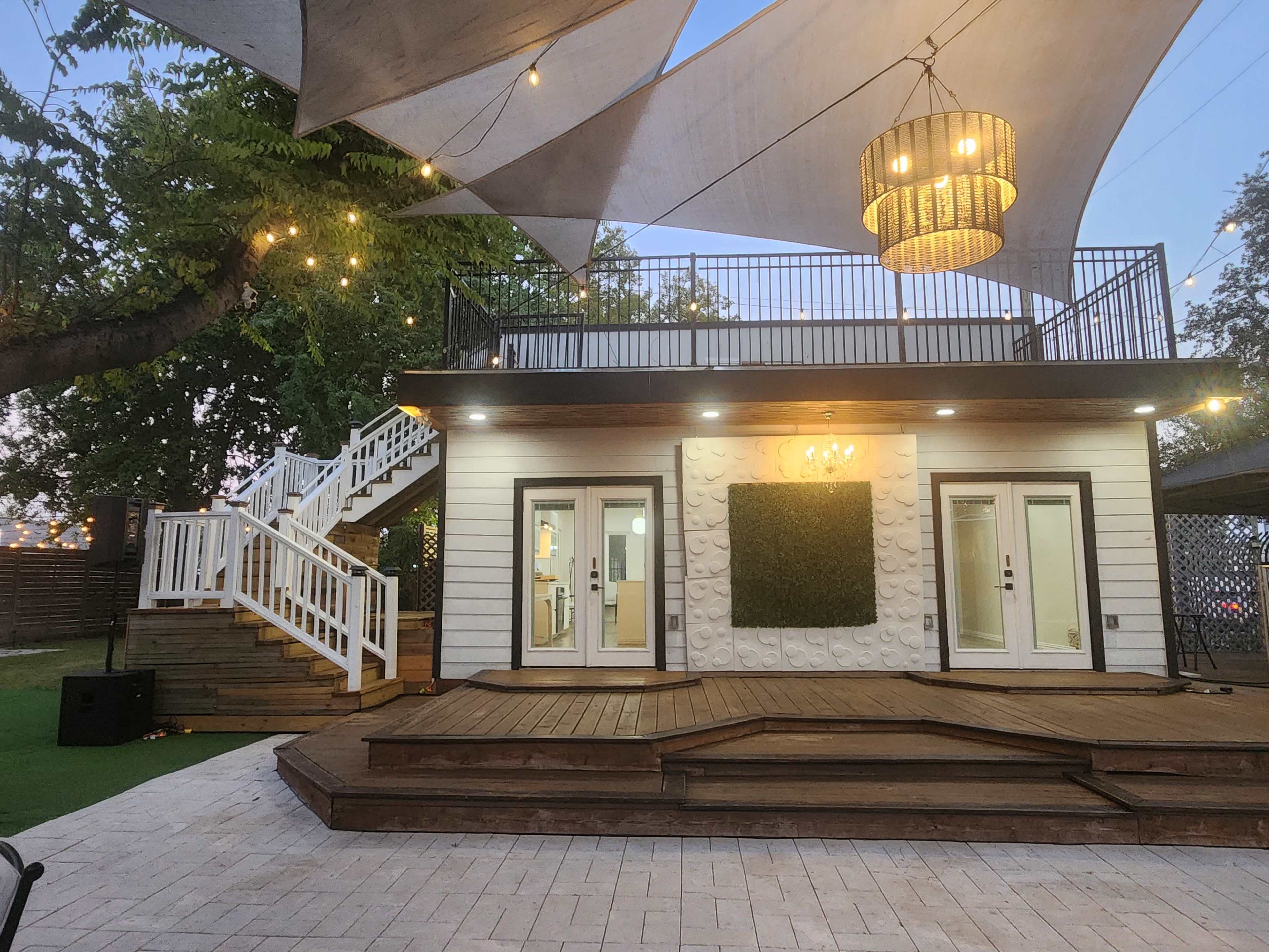 The image shows a modern outdoor area featuring a two-story building with a spacious wooden deck, decorative lighting, and a large canopy overhead.