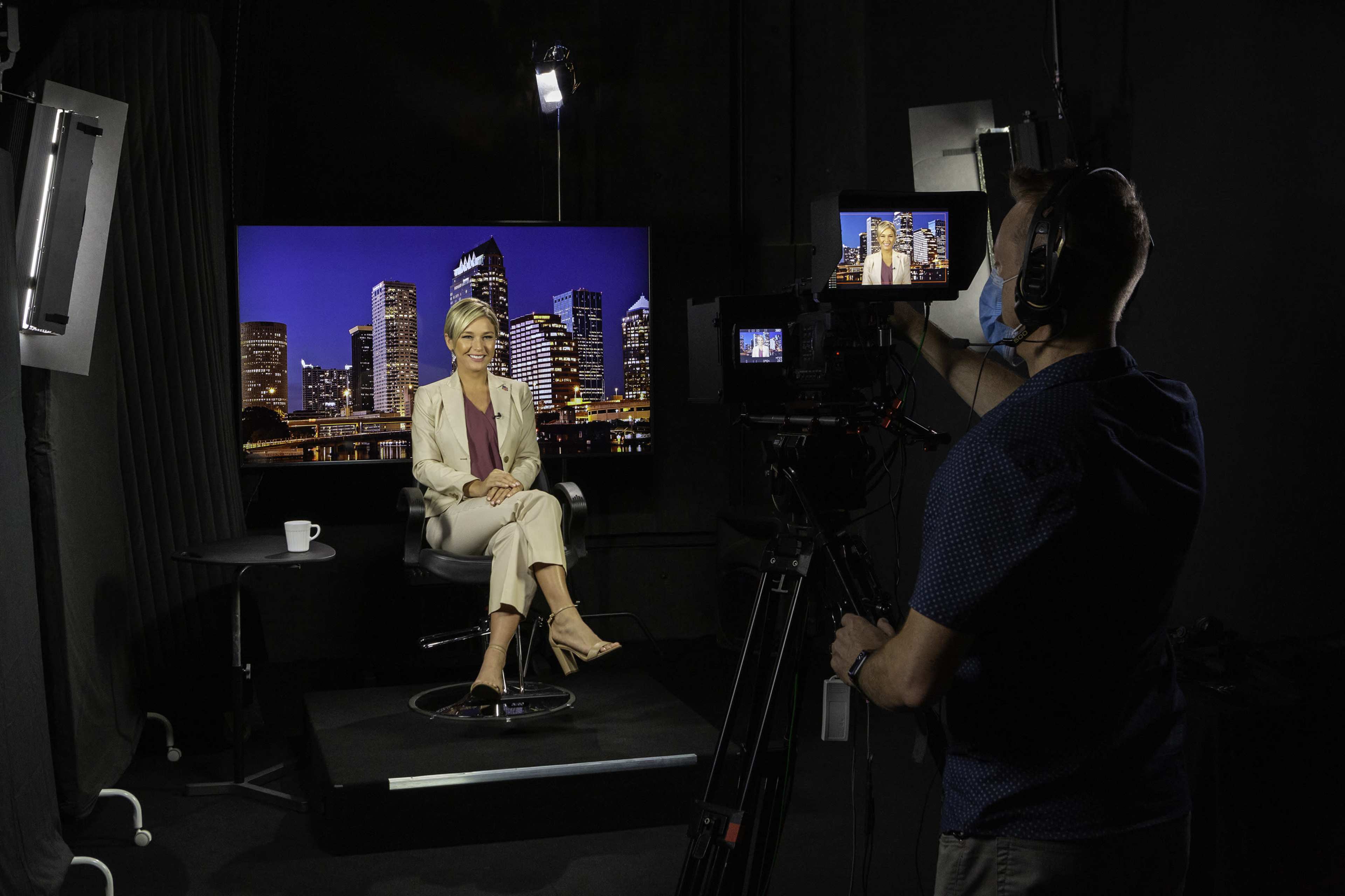 A woman in a light-colored suit is sitting on a chair in a studio, facing a camera while a city skyline is displayed on a monitor behind her.