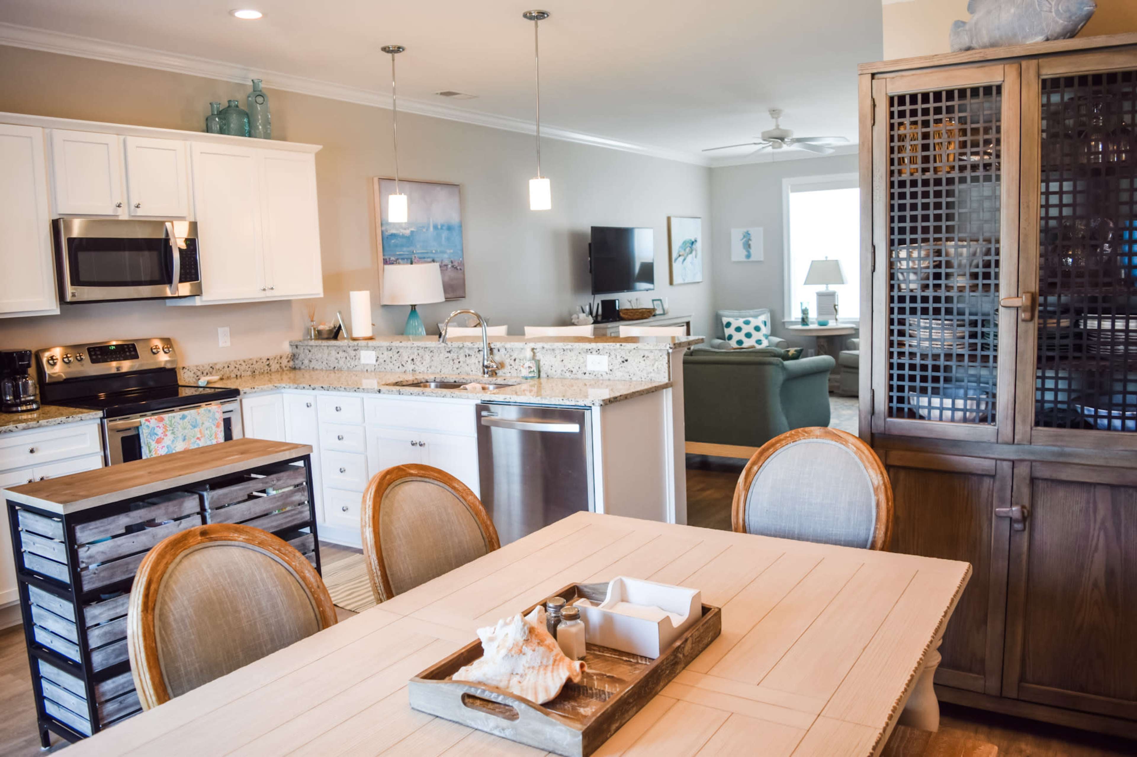 A modern kitchen and dining area features white cabinetry, granite countertops, a wooden dining table with chairs, and a living space with a green sofa in the background.