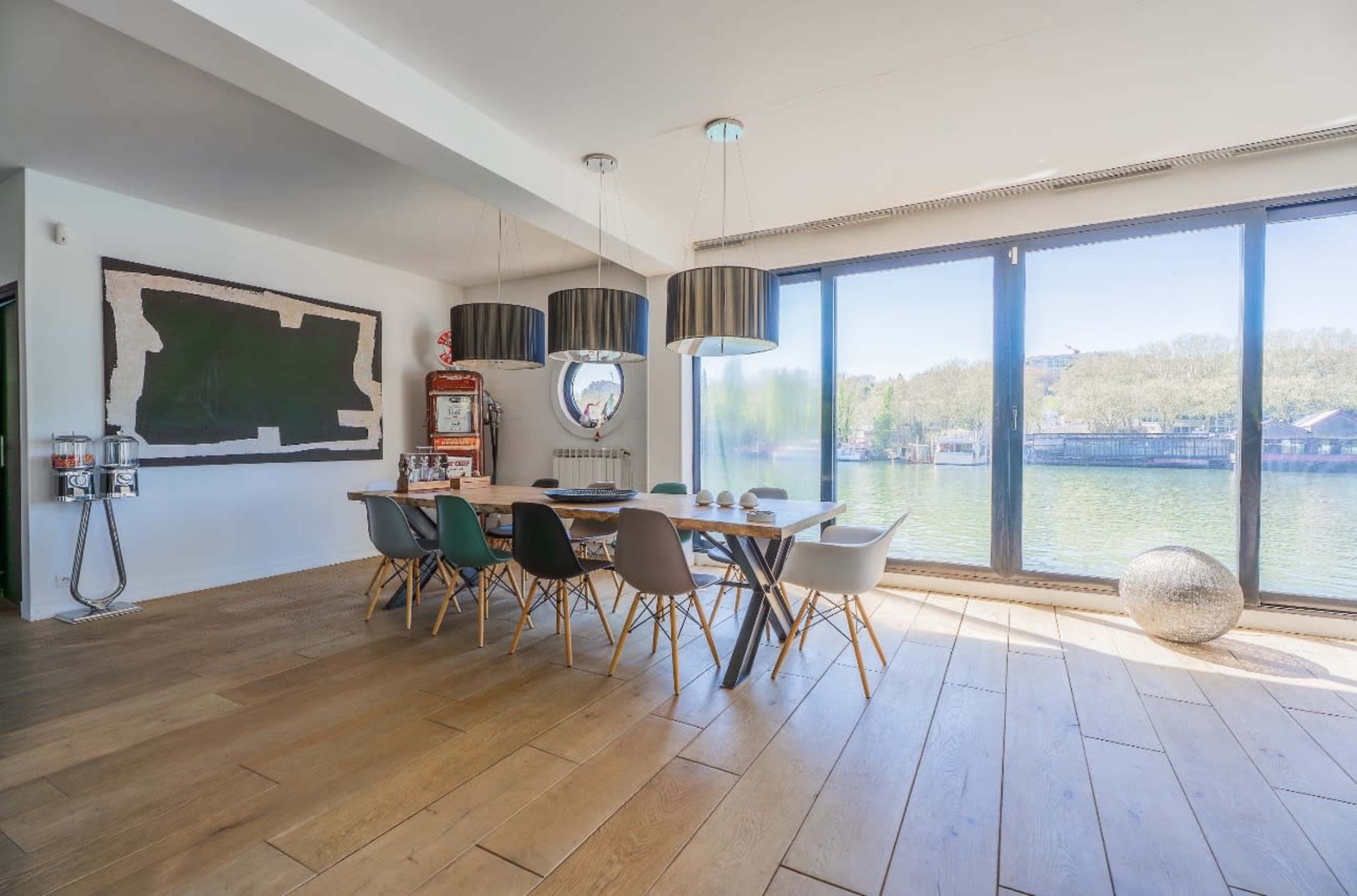 The image shows a modern dining area with a long table surrounded by various chairs, large windows overlooking a body of water, and contemporary light fixtures.