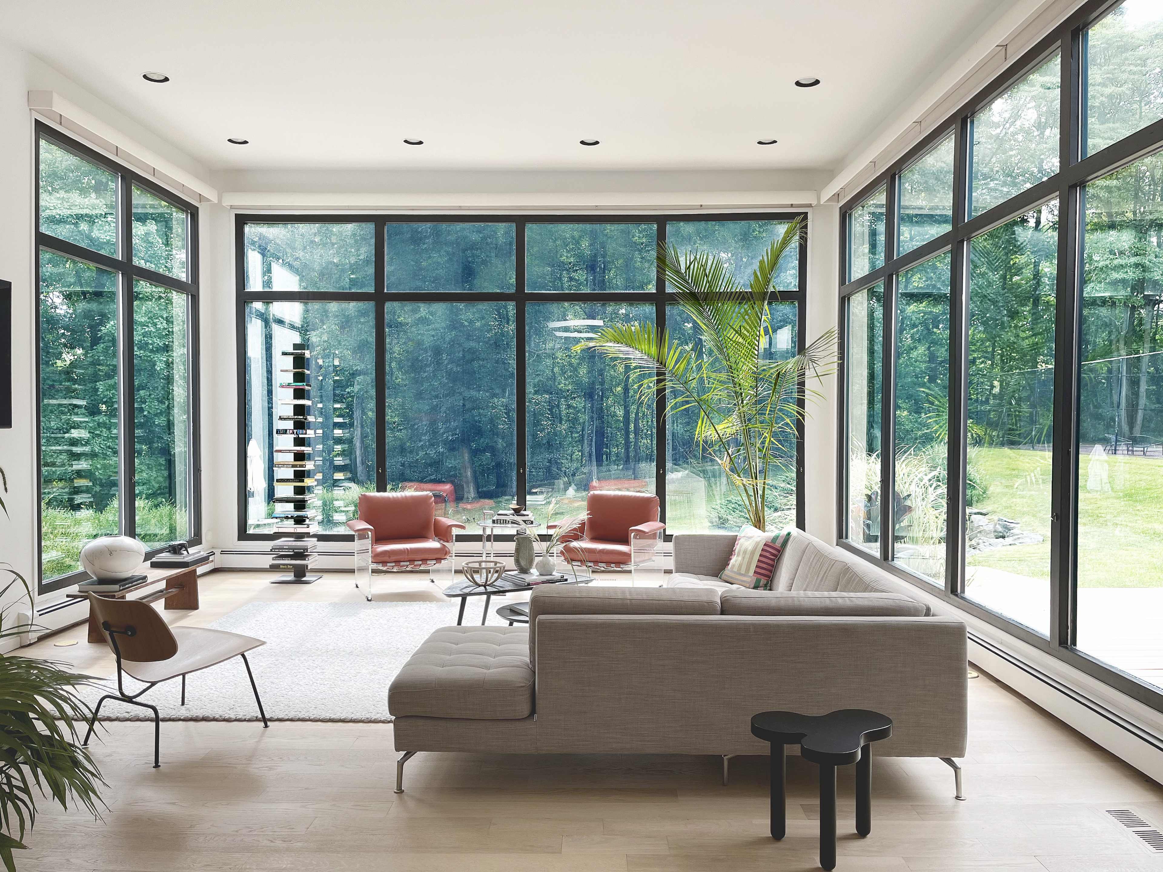 The image depicts a modern living room with large glass windows, minimalist furniture, and greenery visible outside.