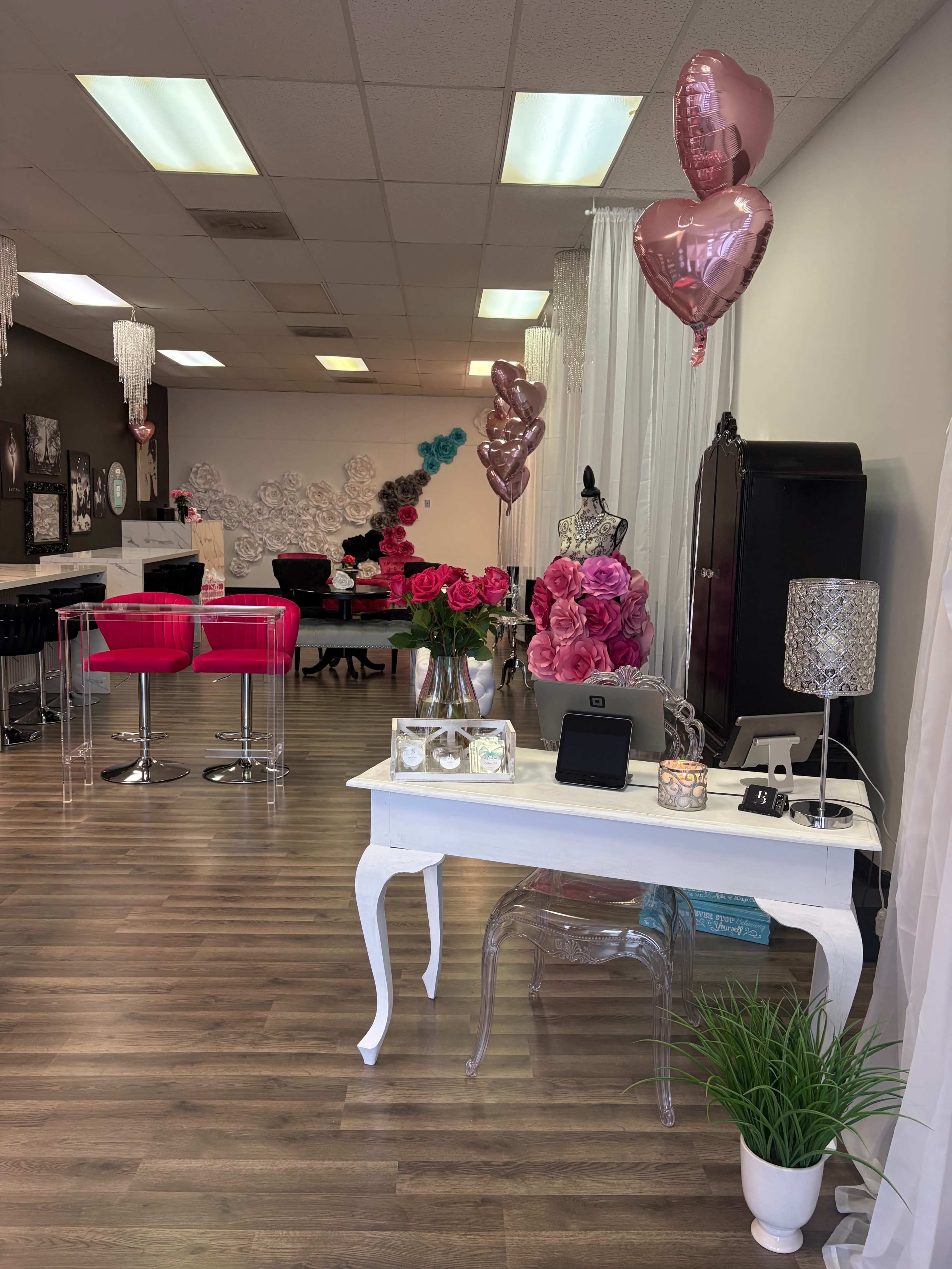 The image shows a brightly decorated interior of a boutique or beauty salon featuring pink seating, floral arrangements, and balloons.