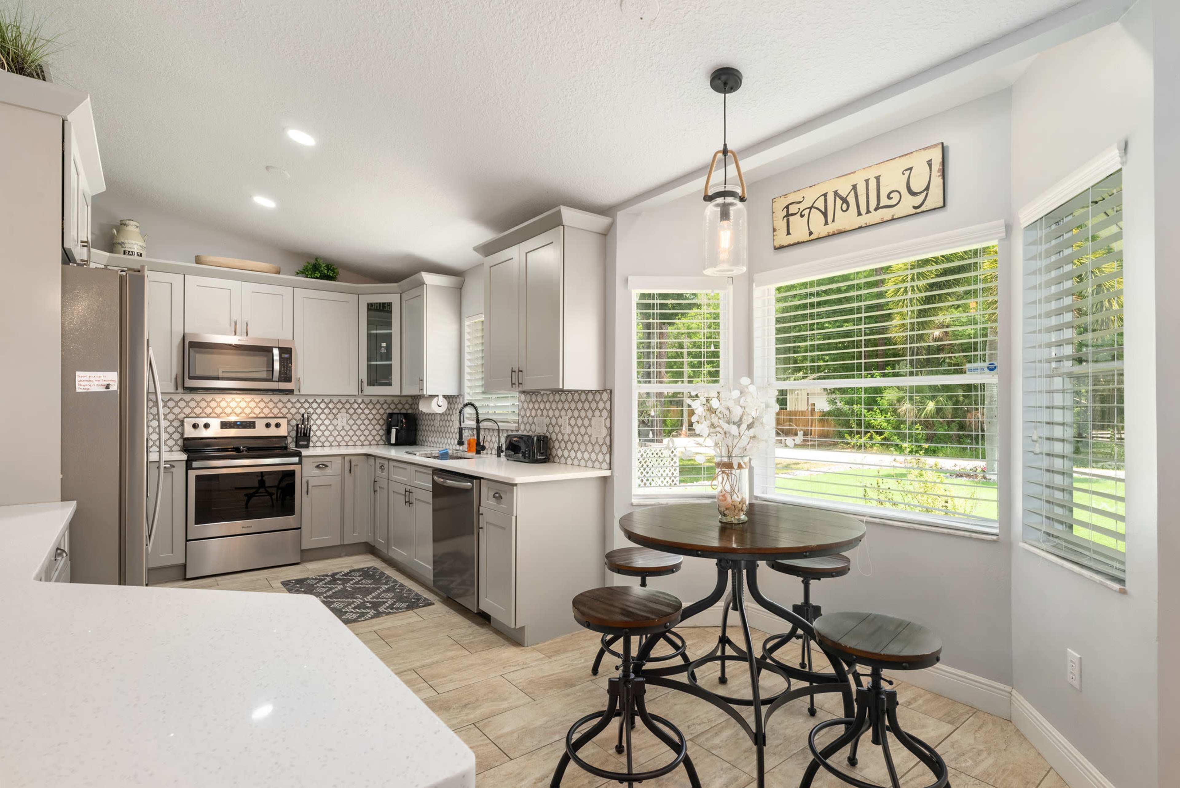 A modern kitchen features gray cabinetry, stainless steel appliances, and a round dining table with four black metal chairs, illuminated by a pendant light.