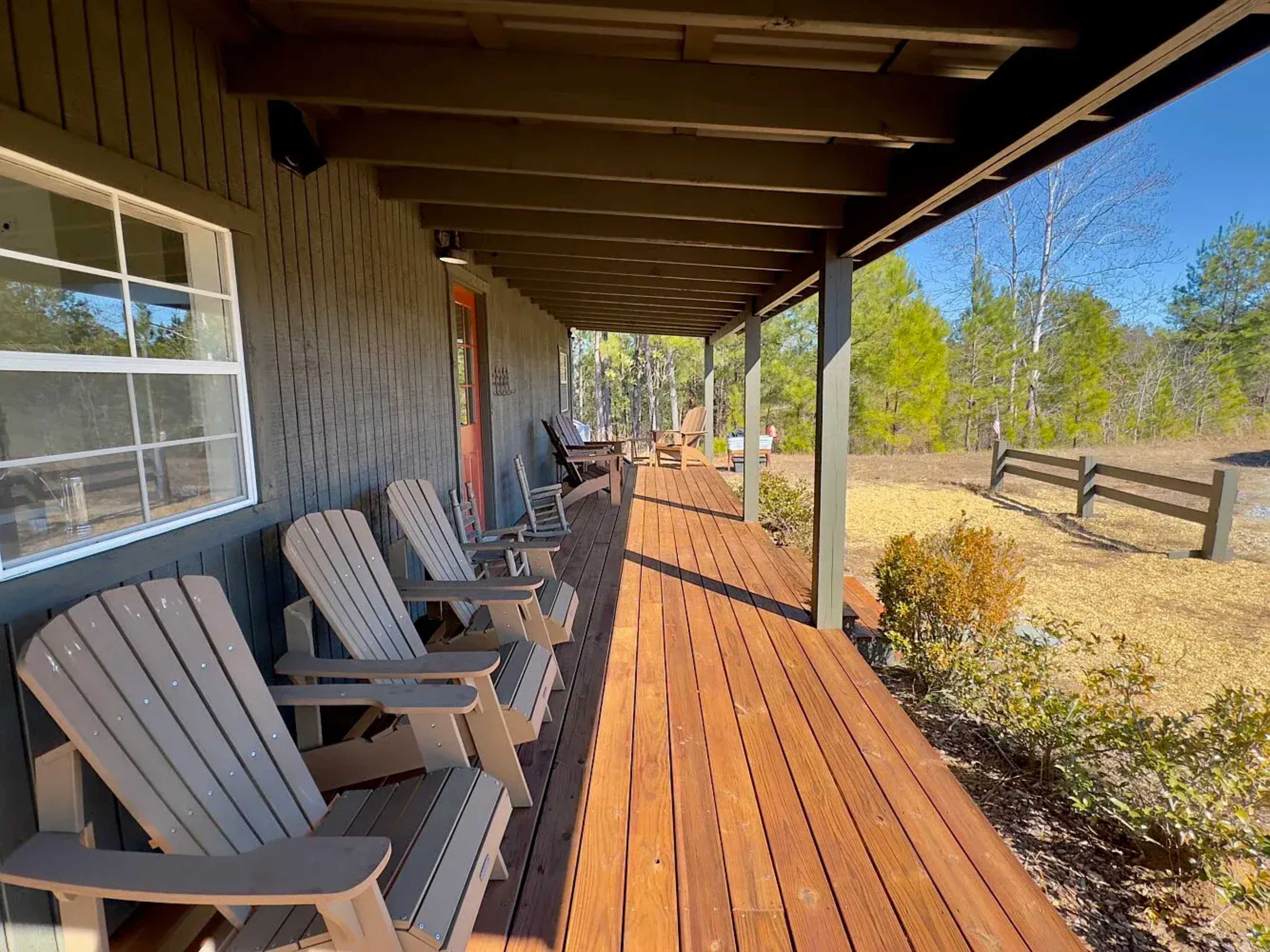 A wooden porch with several chairs overlooks a grassy area and trees in the background.