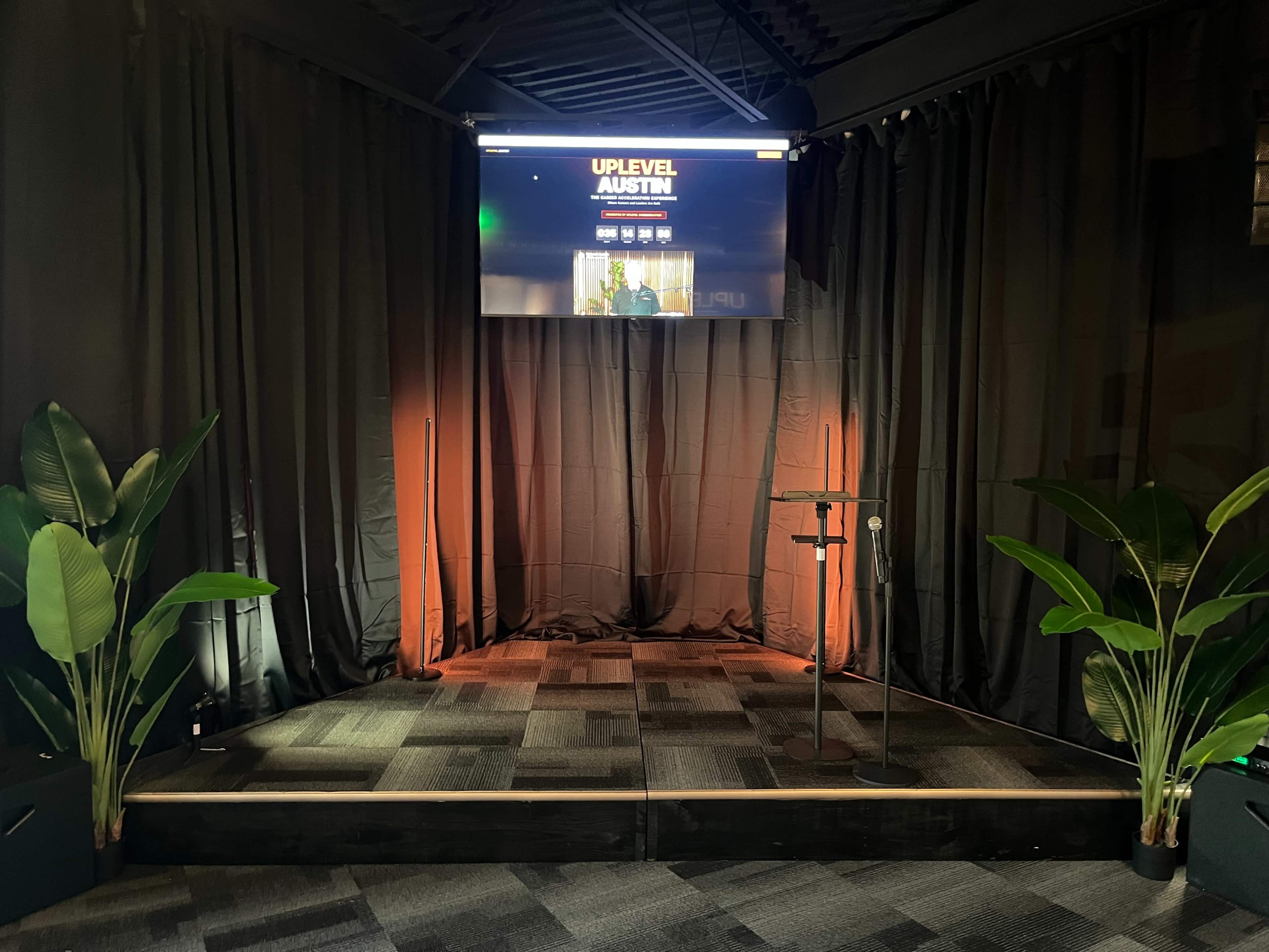 The image shows a stage setup with a microphone stand, surrounded by plants, and a large screen displaying "UPLEVEL AUSTIN" in a dimly lit space.