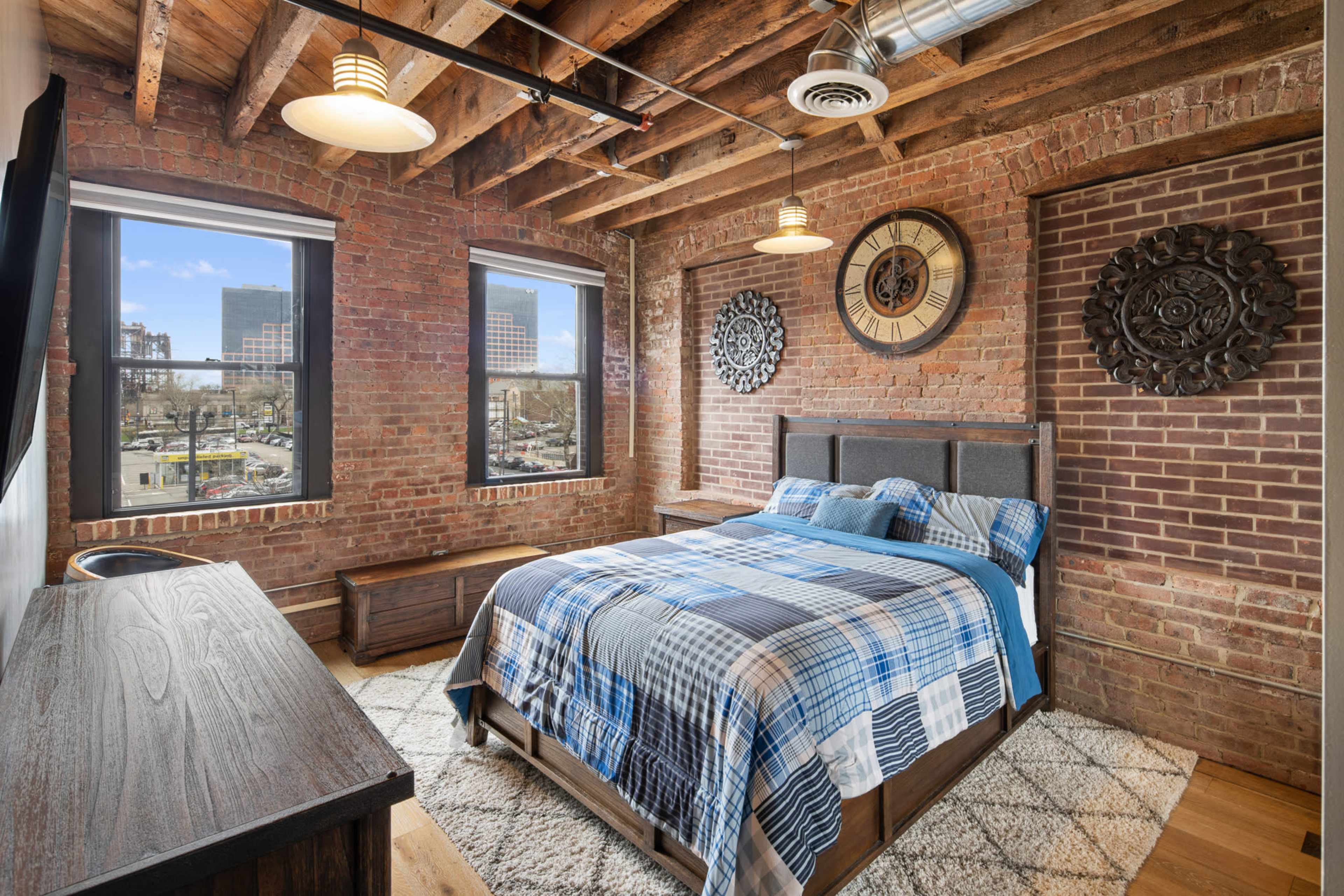 A bedroom features exposed brick walls, large windows overlooking a cityscape, a wooden bed with a blue plaid blanket, and a matching dresser.