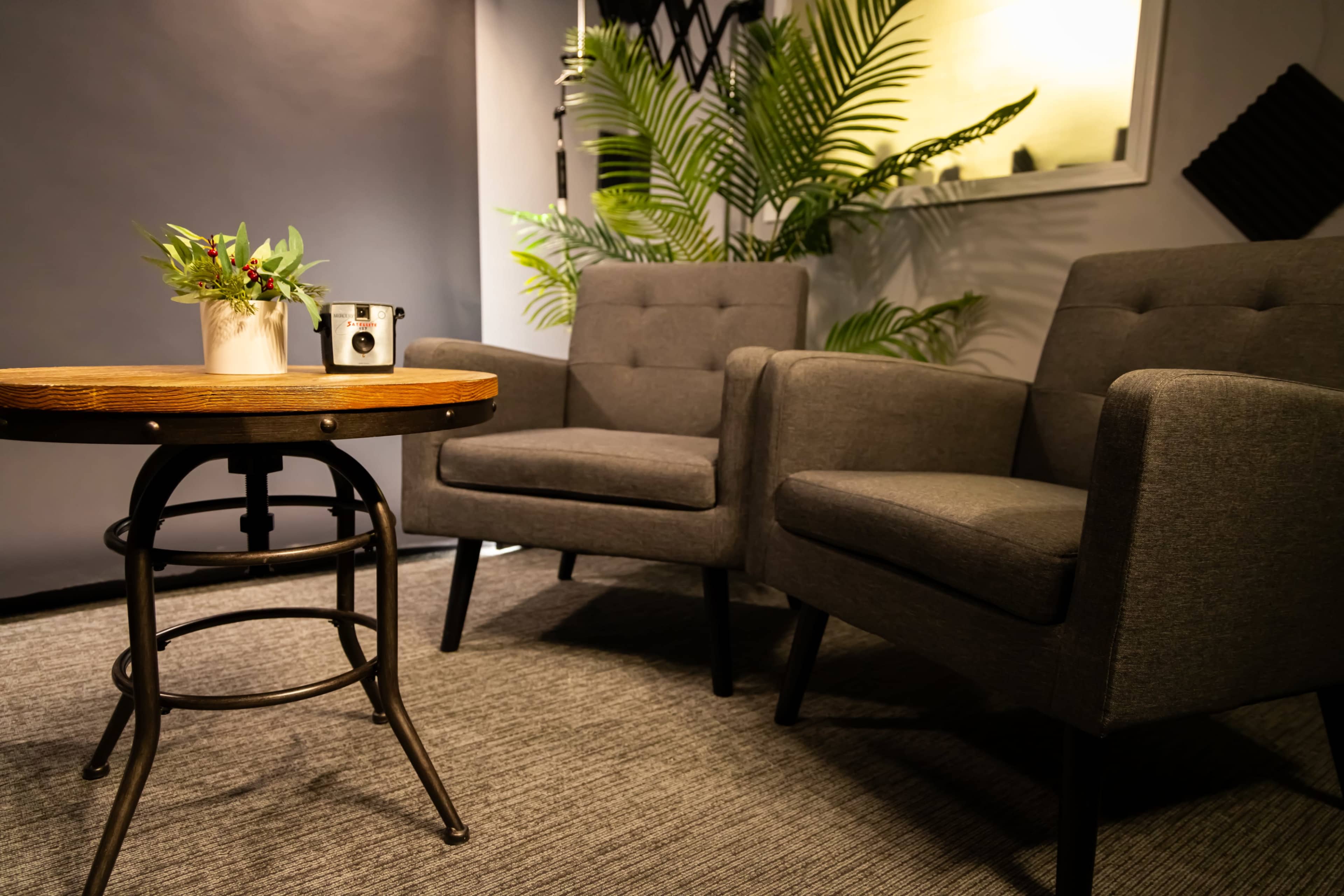 The image depicts a cozy seating area with two gray armchairs and a round wooden table, accompanied by a small plant and a decorative item.